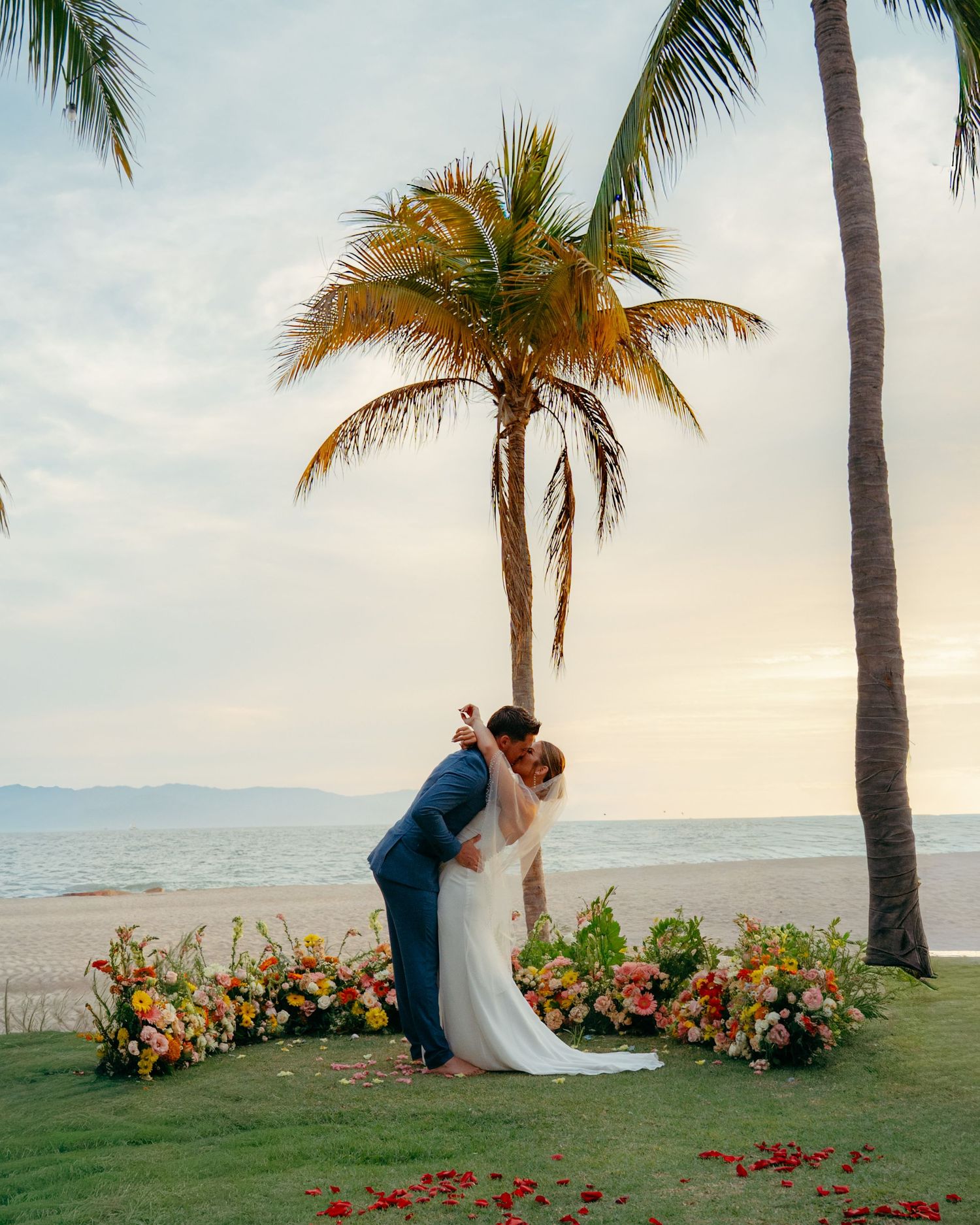 Bride and groom share a first kiss at beachside wedding altar adorned with colorful tropical flowers.