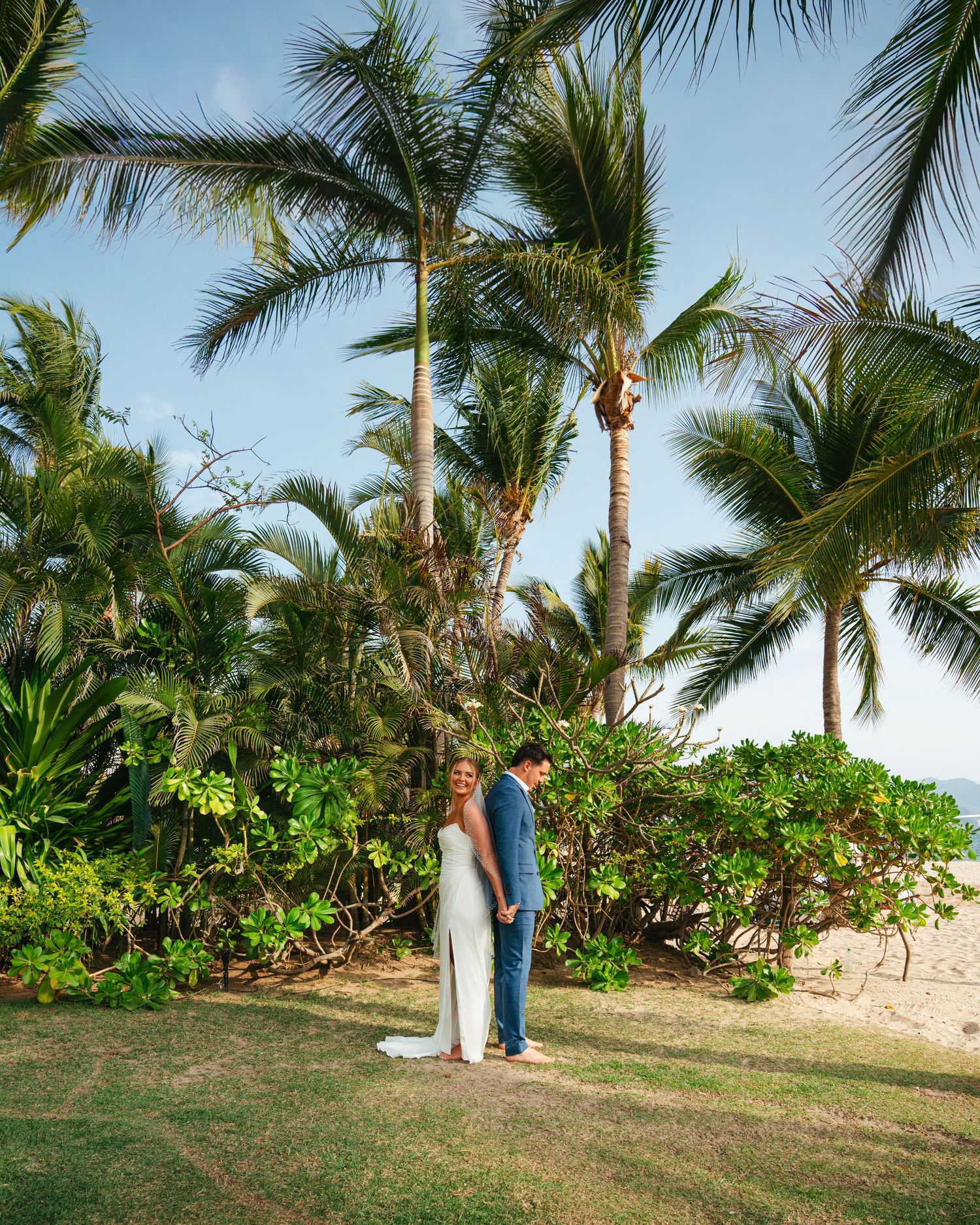 Bride and groom standing together under tall palm trees on a sunny tropical beach wedding.