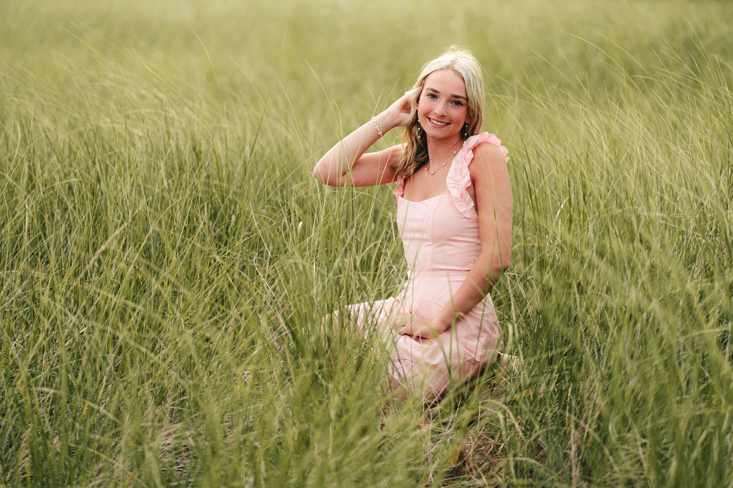 Smiling blonde woman in pink ruffled dress kneels in tall green grass field.
