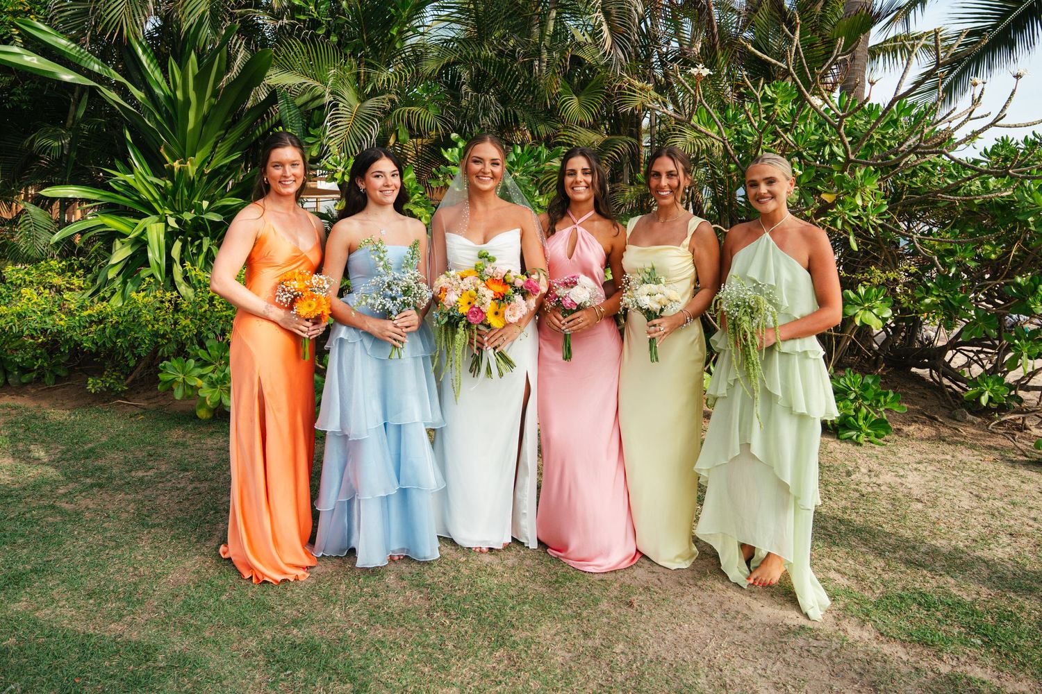 Six bridesmaids in colorful mismatched dresses holding bouquets in a tropical garden setting.