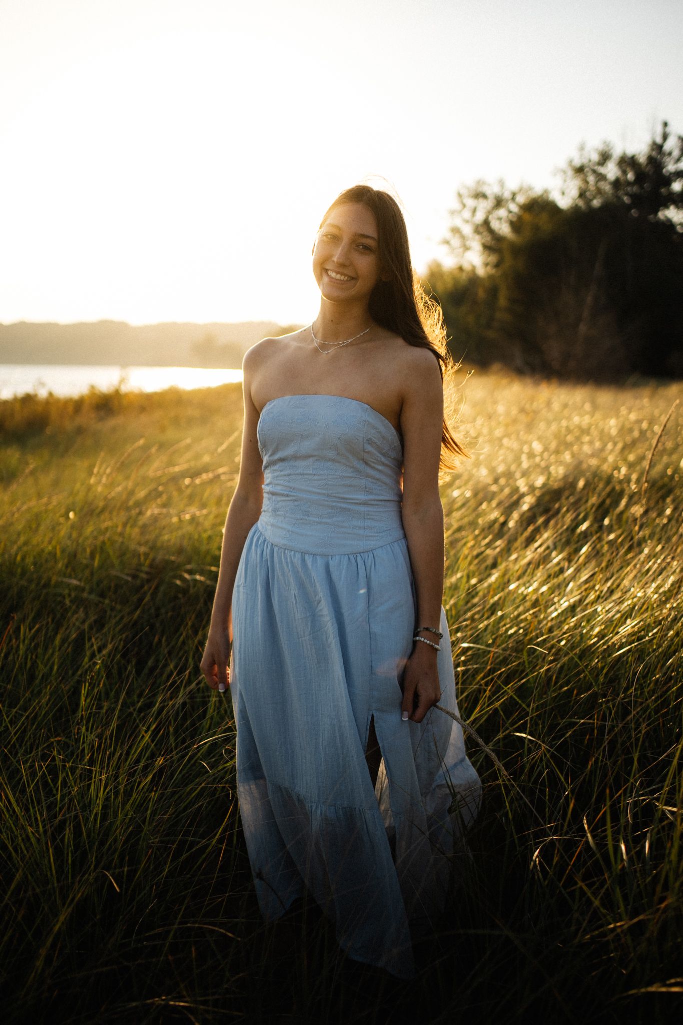 Young woman in a light blue strapless dress smiling in a golden sunset field near a lake.