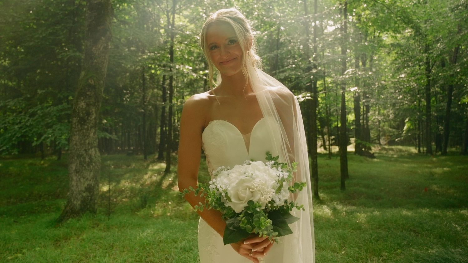A blonde bride in a white gown holds a bouquet of white flowers in a sunlit forest setting.