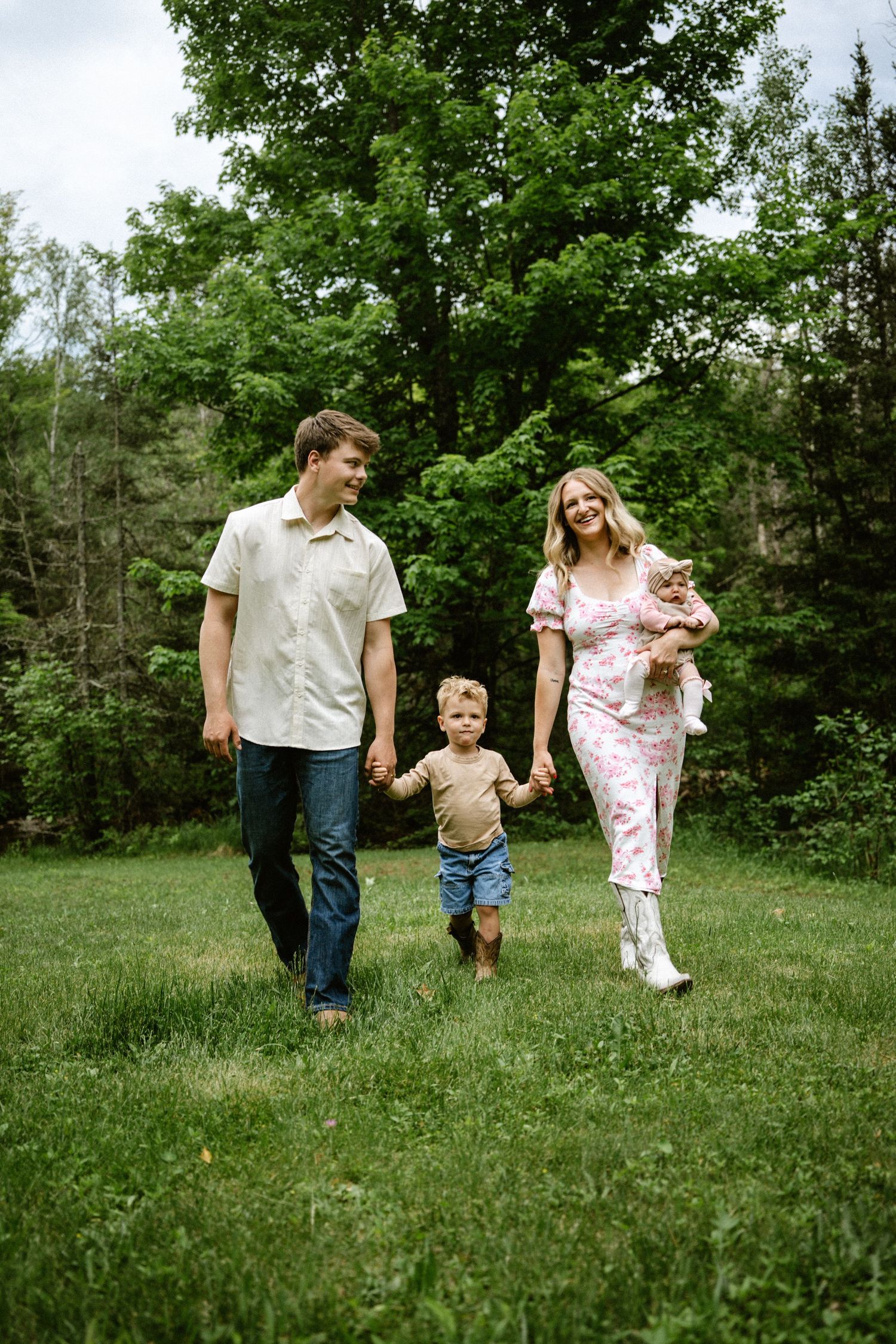 A happy family of three walks hand-in-hand through a lush green park, with parents holding a toddler and baby outdoors.