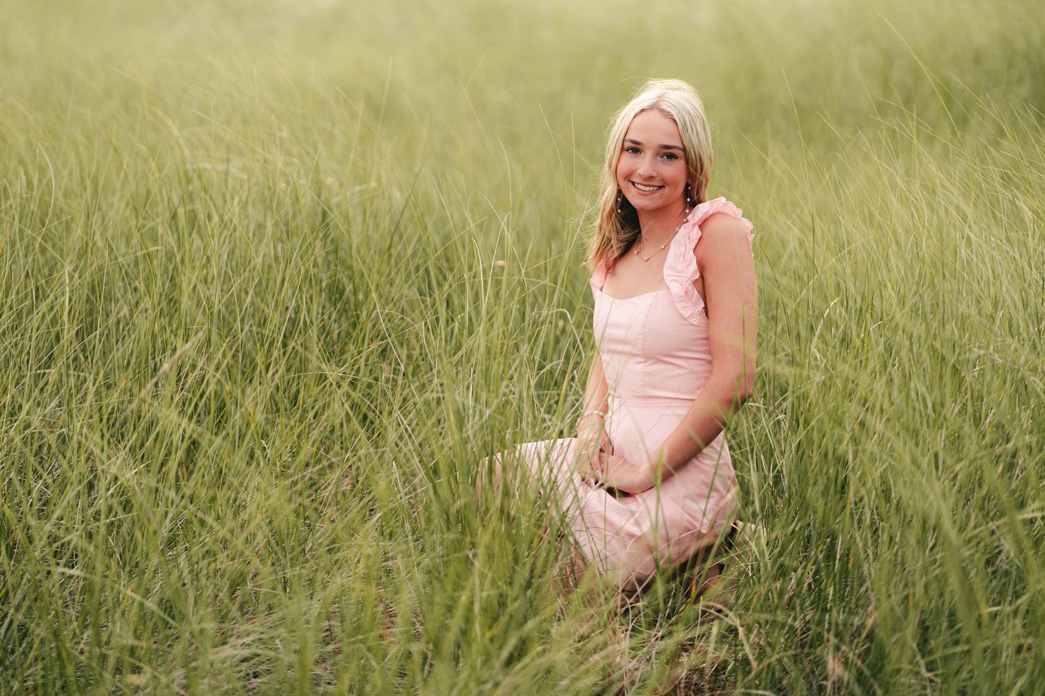 Blonde young woman in pink dress kneeling in a tall green grass field senior portrait.