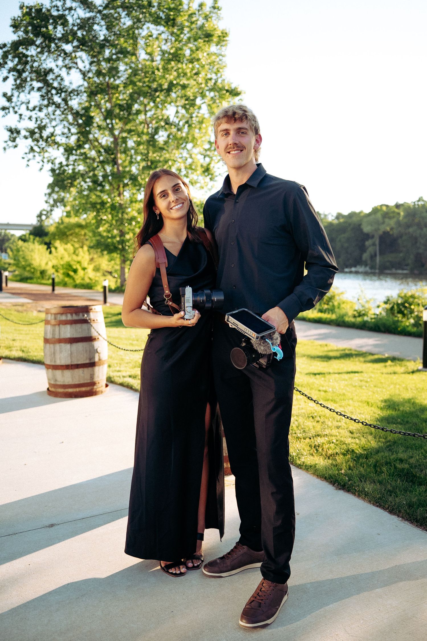 A couple dressed in black formal attire poses outdoors near a waterfront, holding cameras on a sunny day.