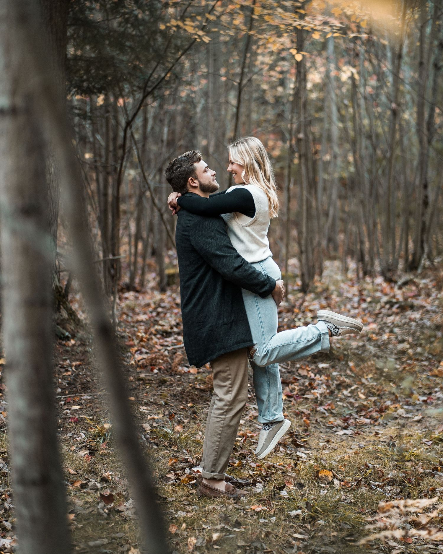 Couple sharing joyful embrace in fall woodland, man lifting woman among colorful leaves.