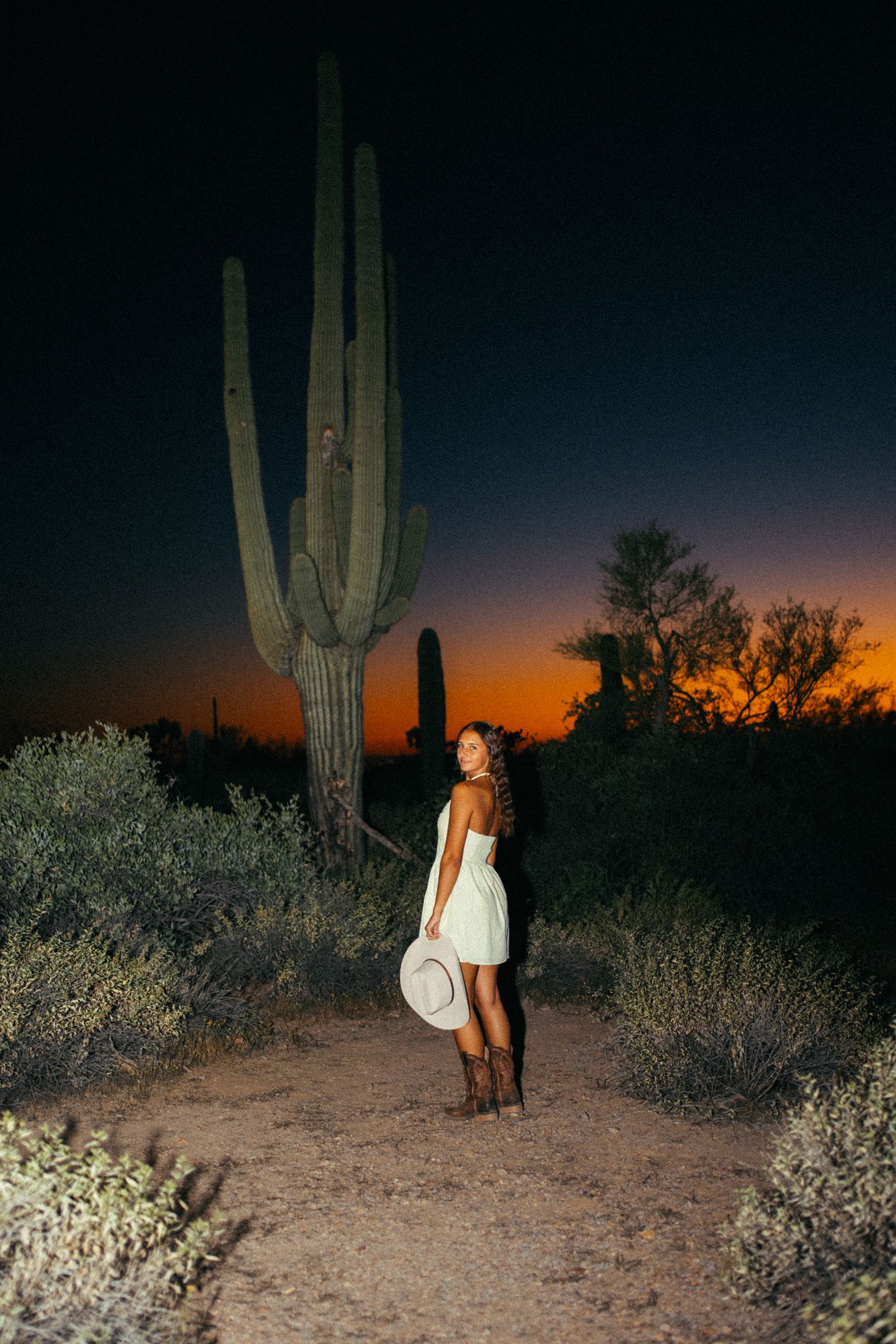 Woman in white dress and cowboy boots poses beside tall saguaro cactus in Arizona desert at sunset.