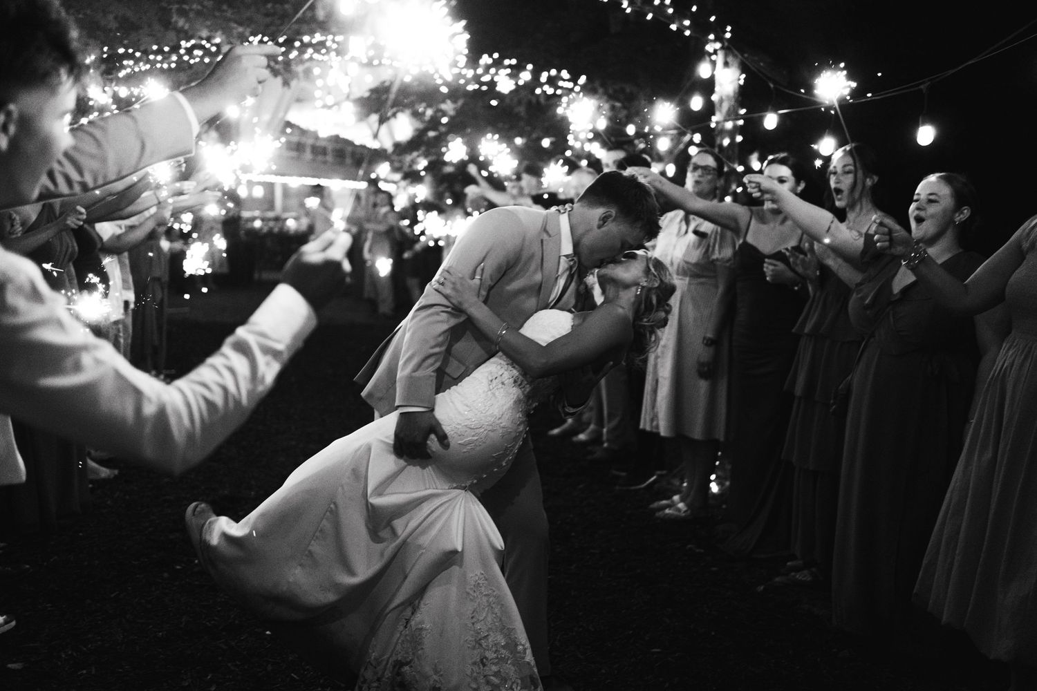 Groom lifts bride during sparkler exit in dramatic black and white wedding night celebration photo.
