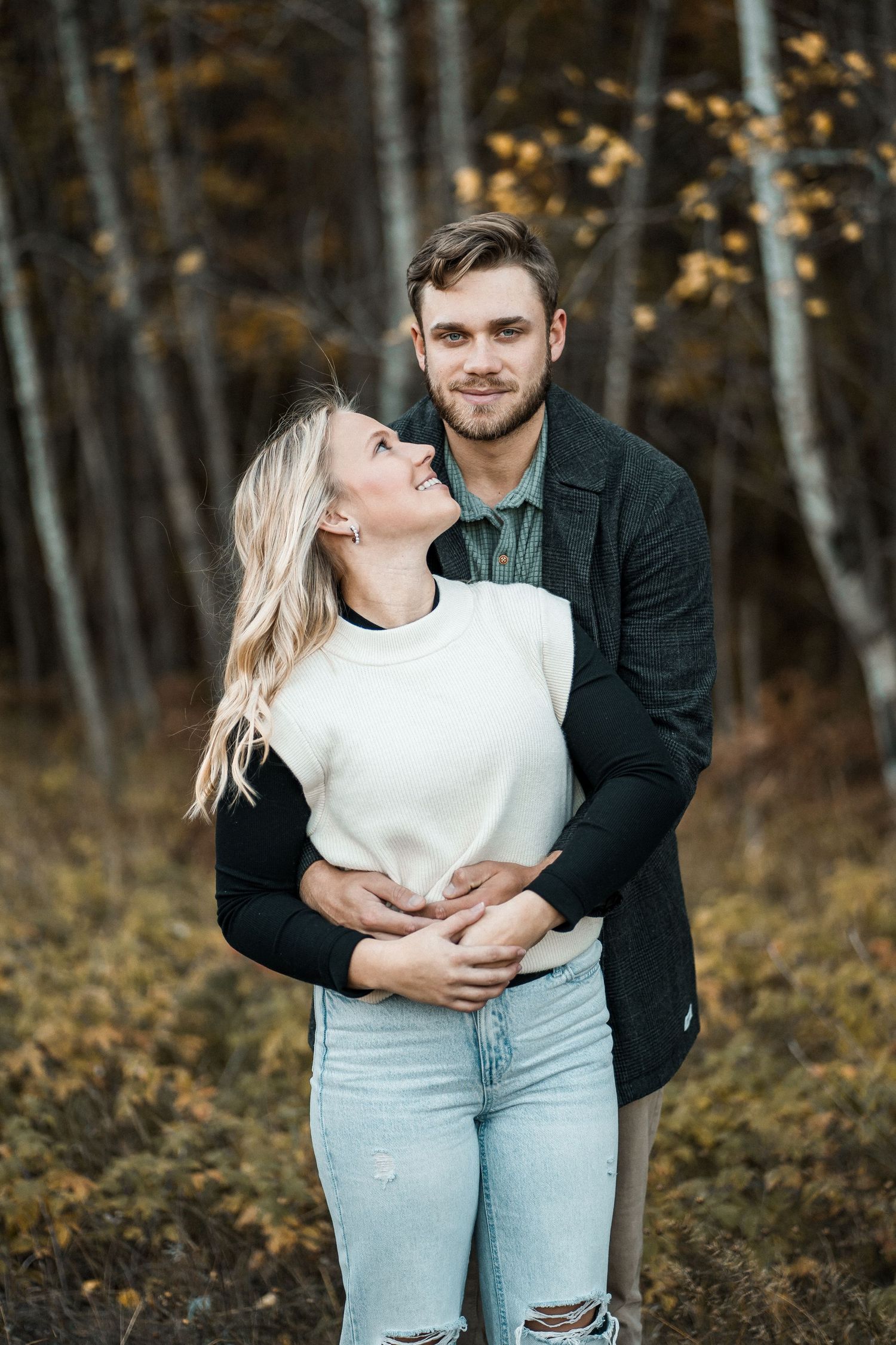 Couple embracing in autumn forest, woman in white sweater laughing up at smiling man.