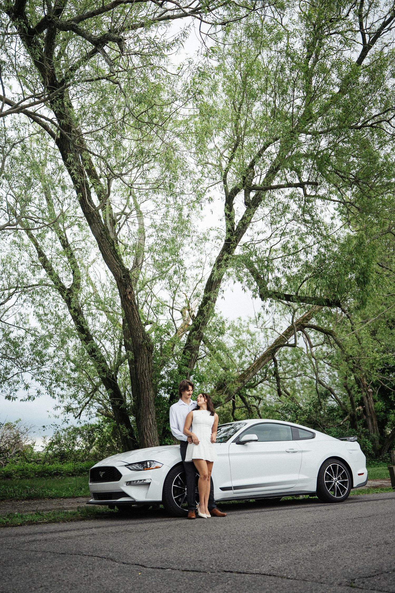 Couple in white outfits posing by a white Ford Mustang under large green trees on a paved road.