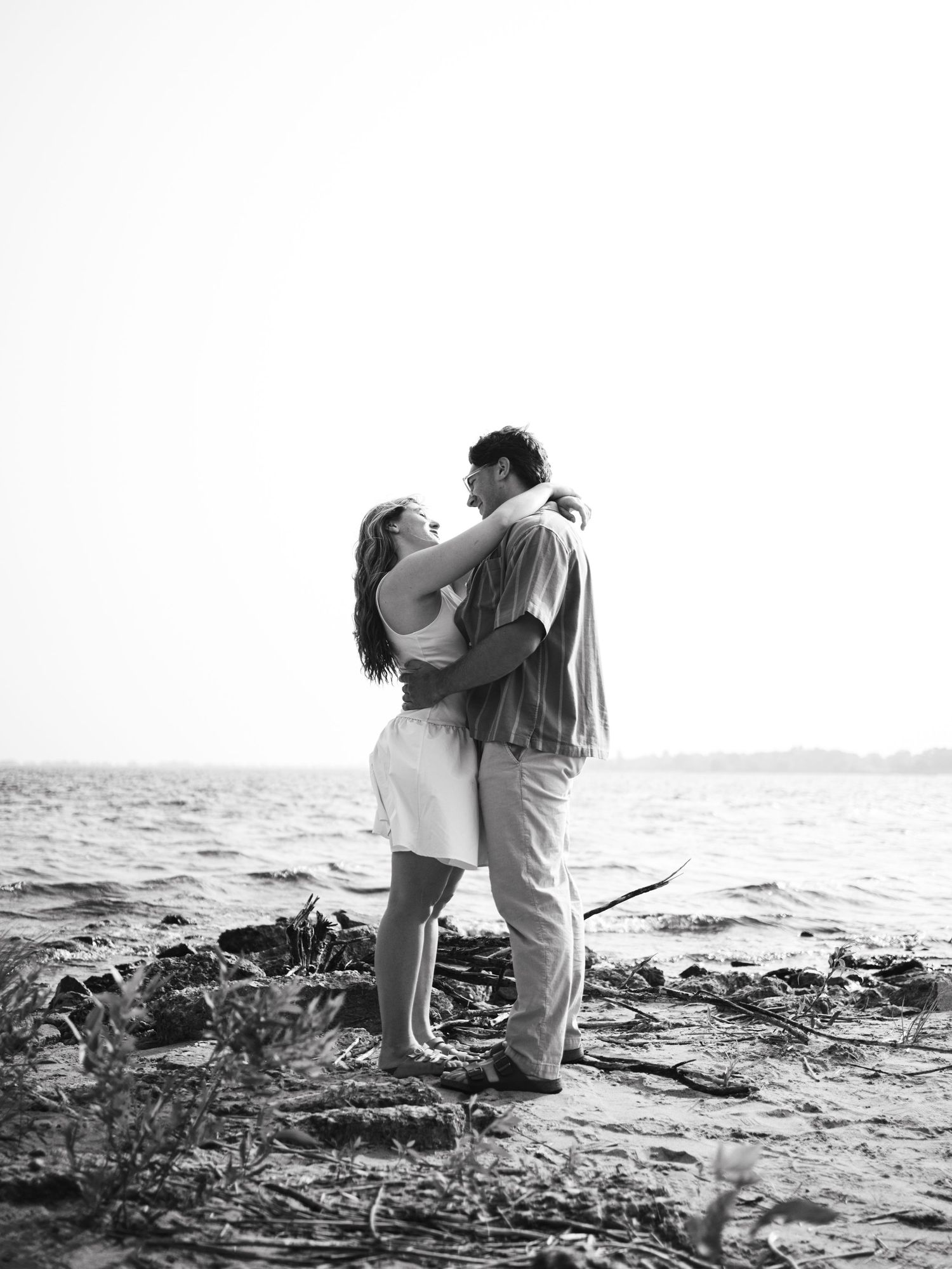 Black and white photo of a romantic couple embracing on a rocky shoreline with water in the background.