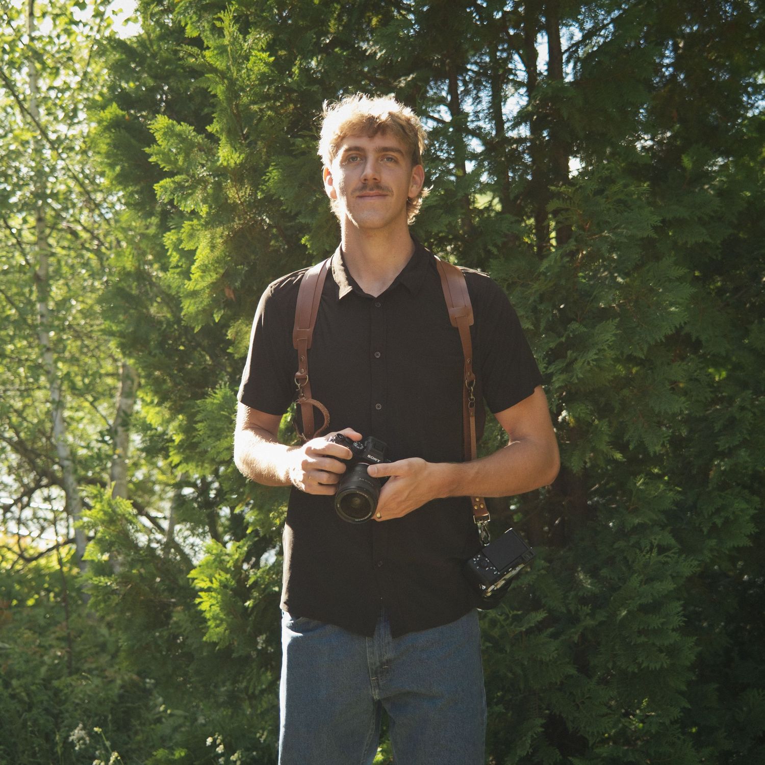 Young man with curly hair holding a camera outdoors, wearing a black shirt with leather straps, surrounded by green trees.