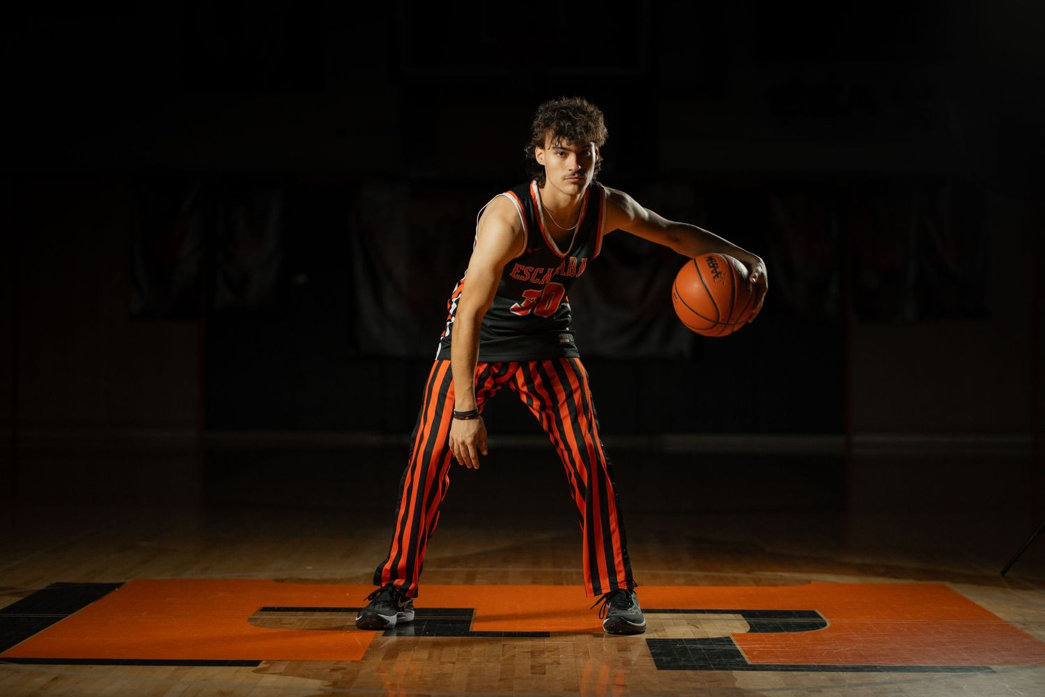 Teen basketball player in red and black uniform dribbling on a dark dramatic indoor court.