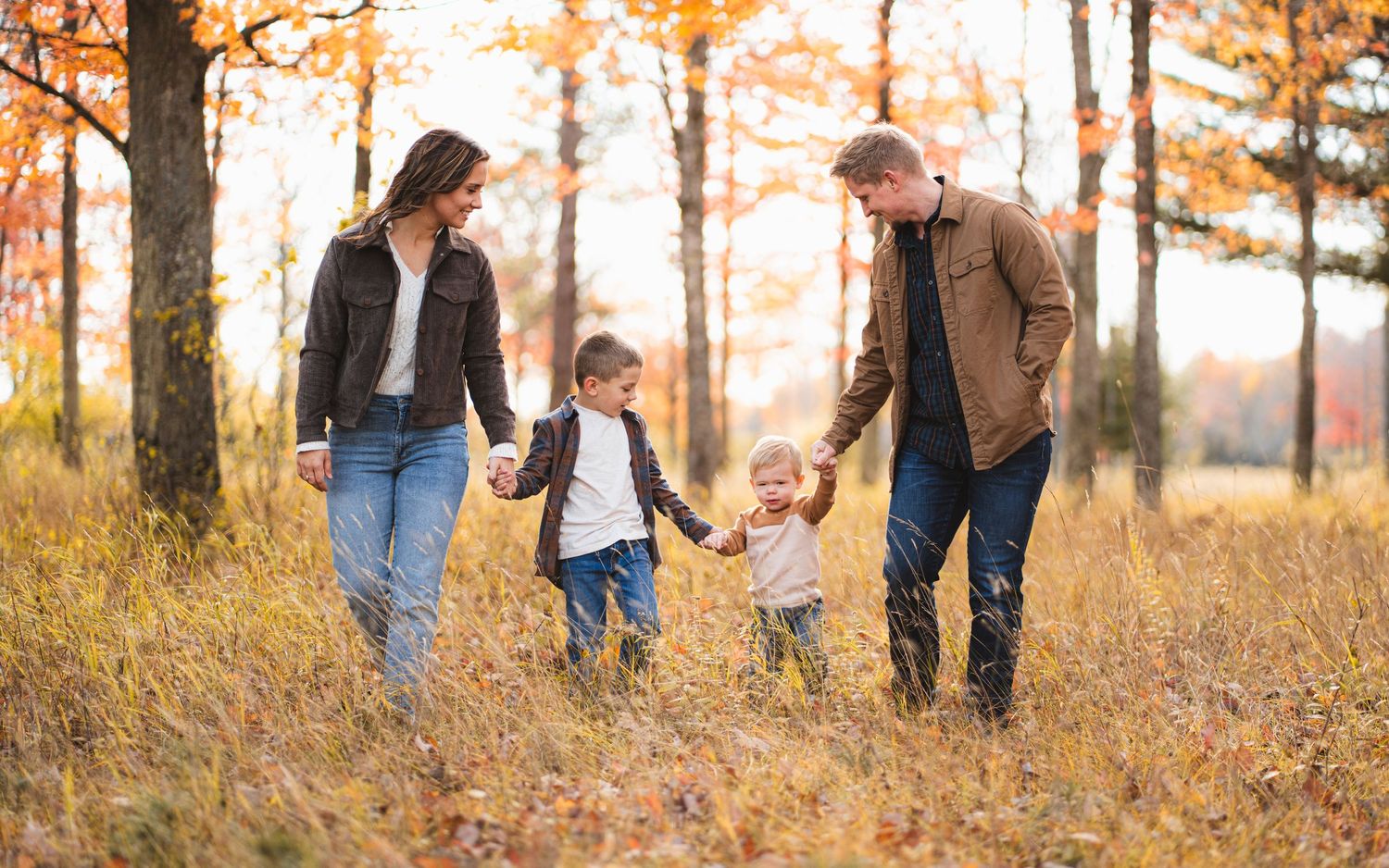 Happy family of four walking hand-in-hand through a golden autumn forest with colorful fall foliage.