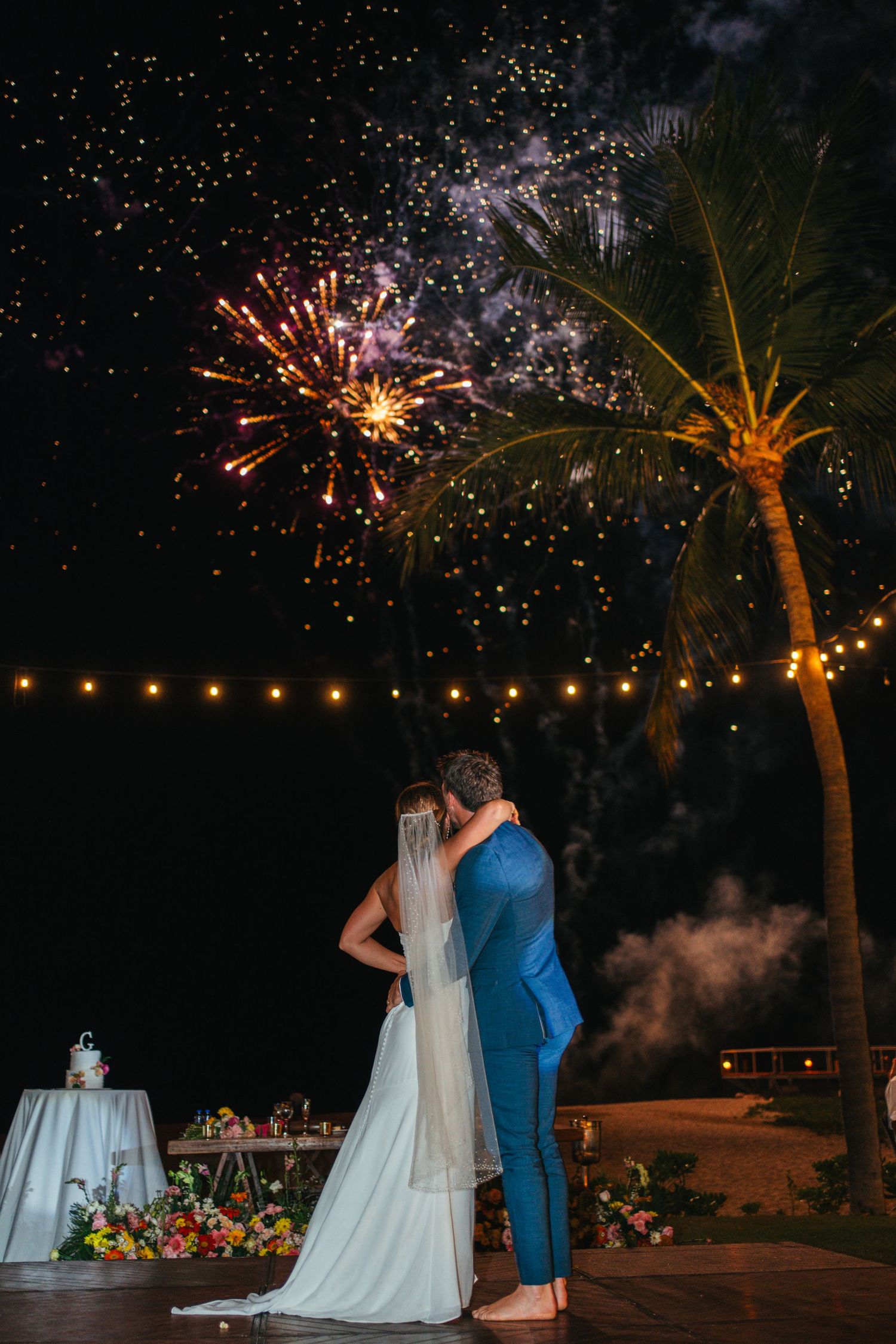 Bride and groom watch stunning fireworks display together at night during tropical beach wedding reception.