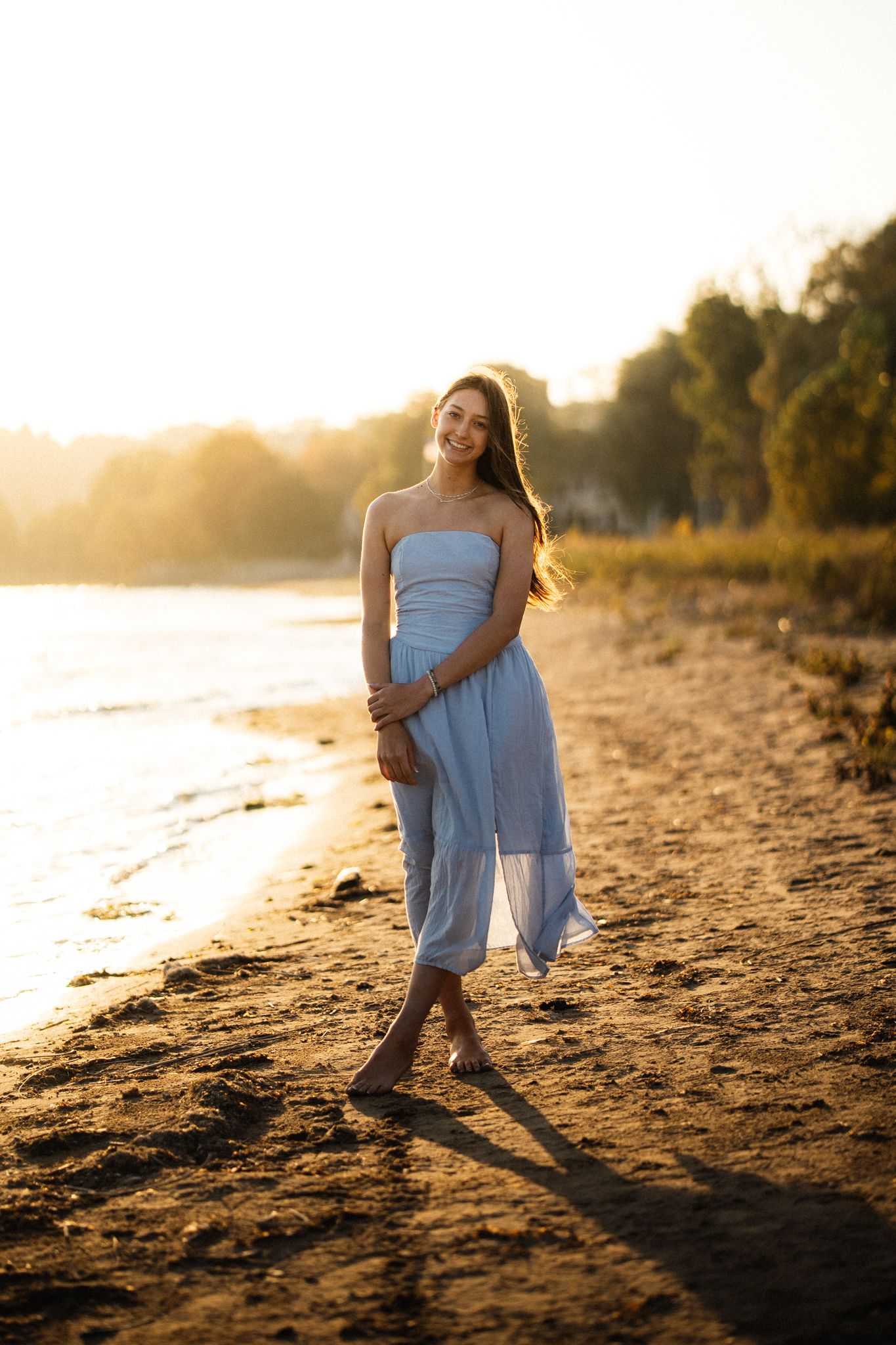 Young woman in light blue strapless dress walking barefoot on sandy beach at golden sunset.