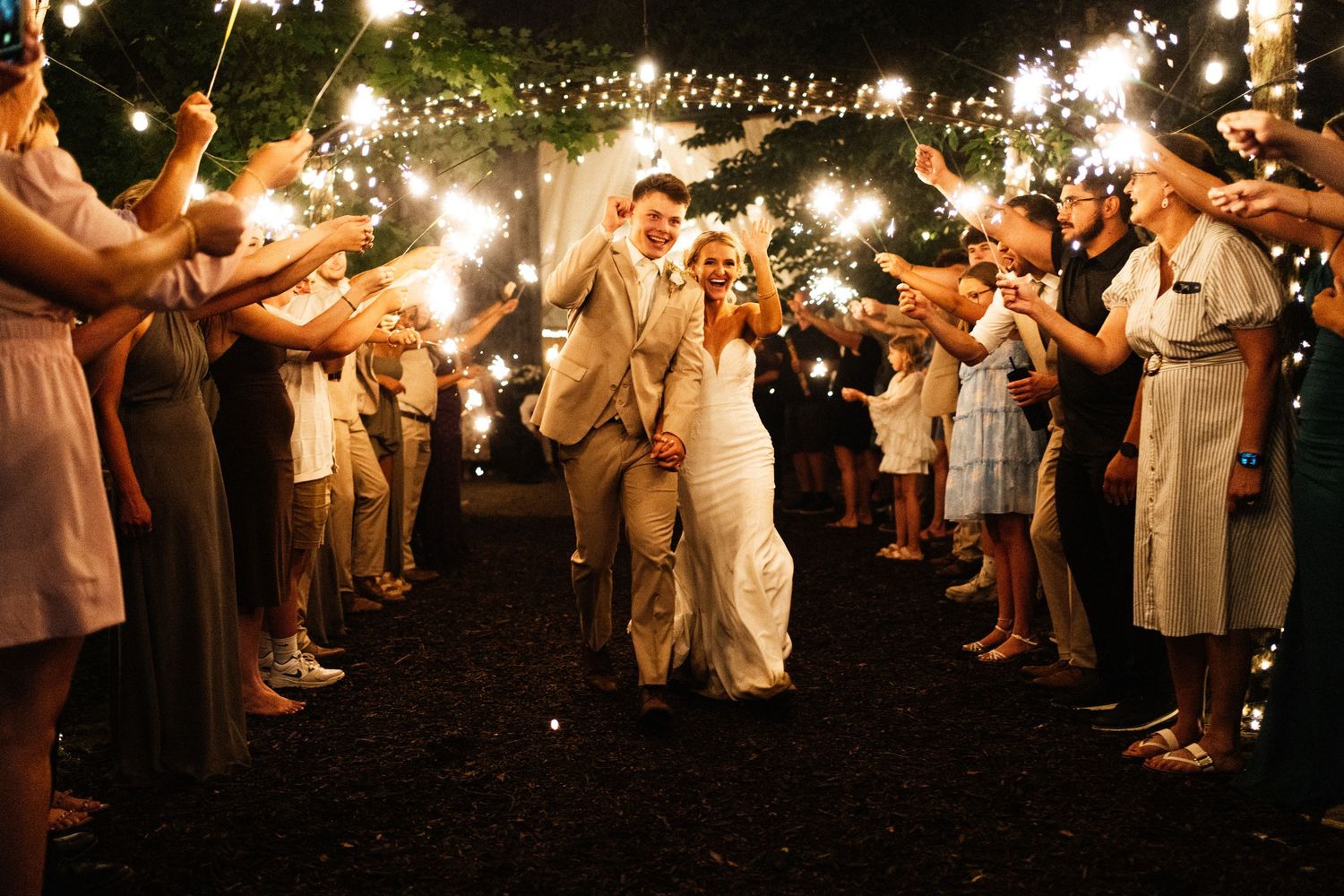 Bride and groom celebrating their sparkler exit surrounded by cheering guests at their evening wedding.