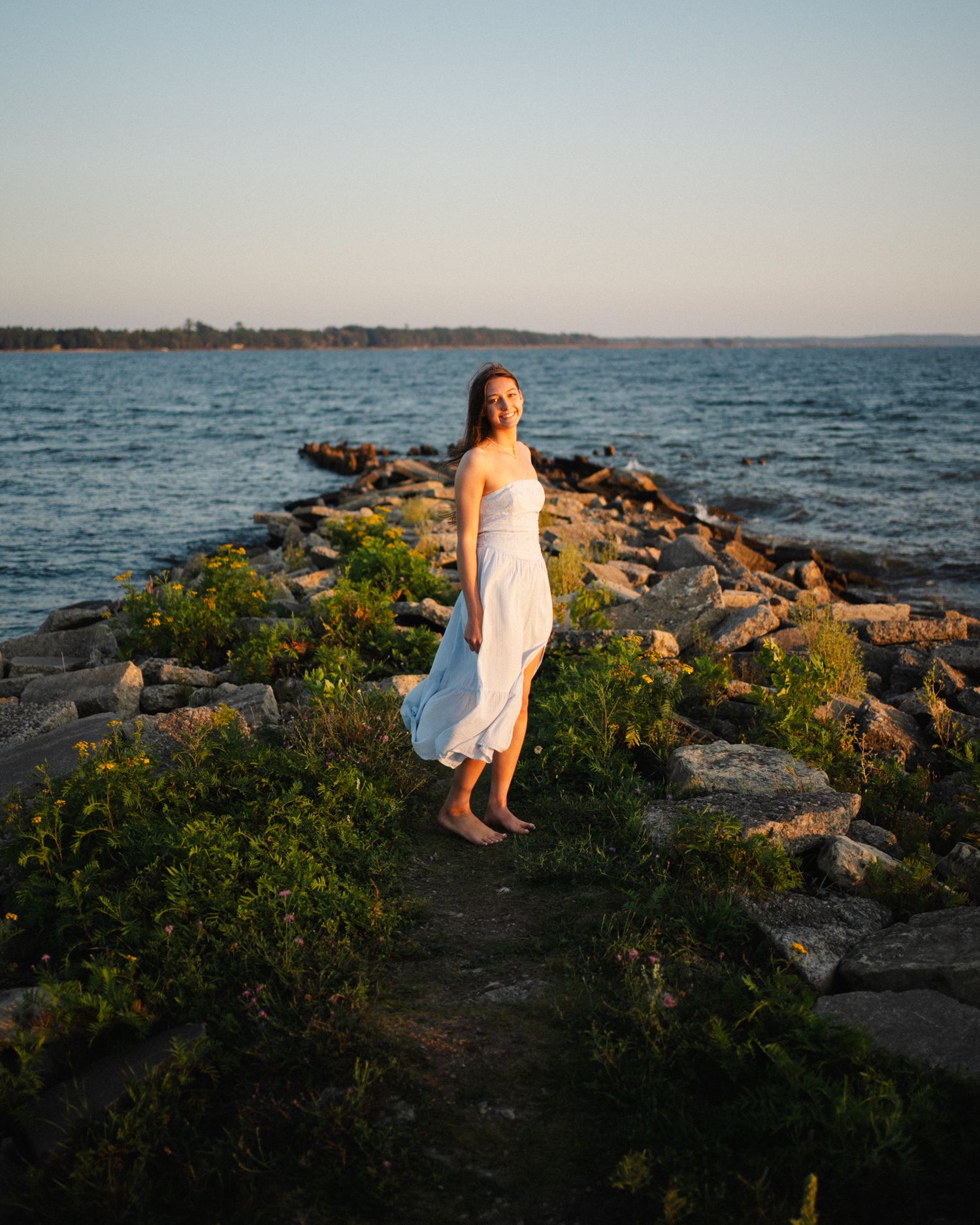 Young woman in white strapless dress standing on rocky shoreline at golden hour by the sea.