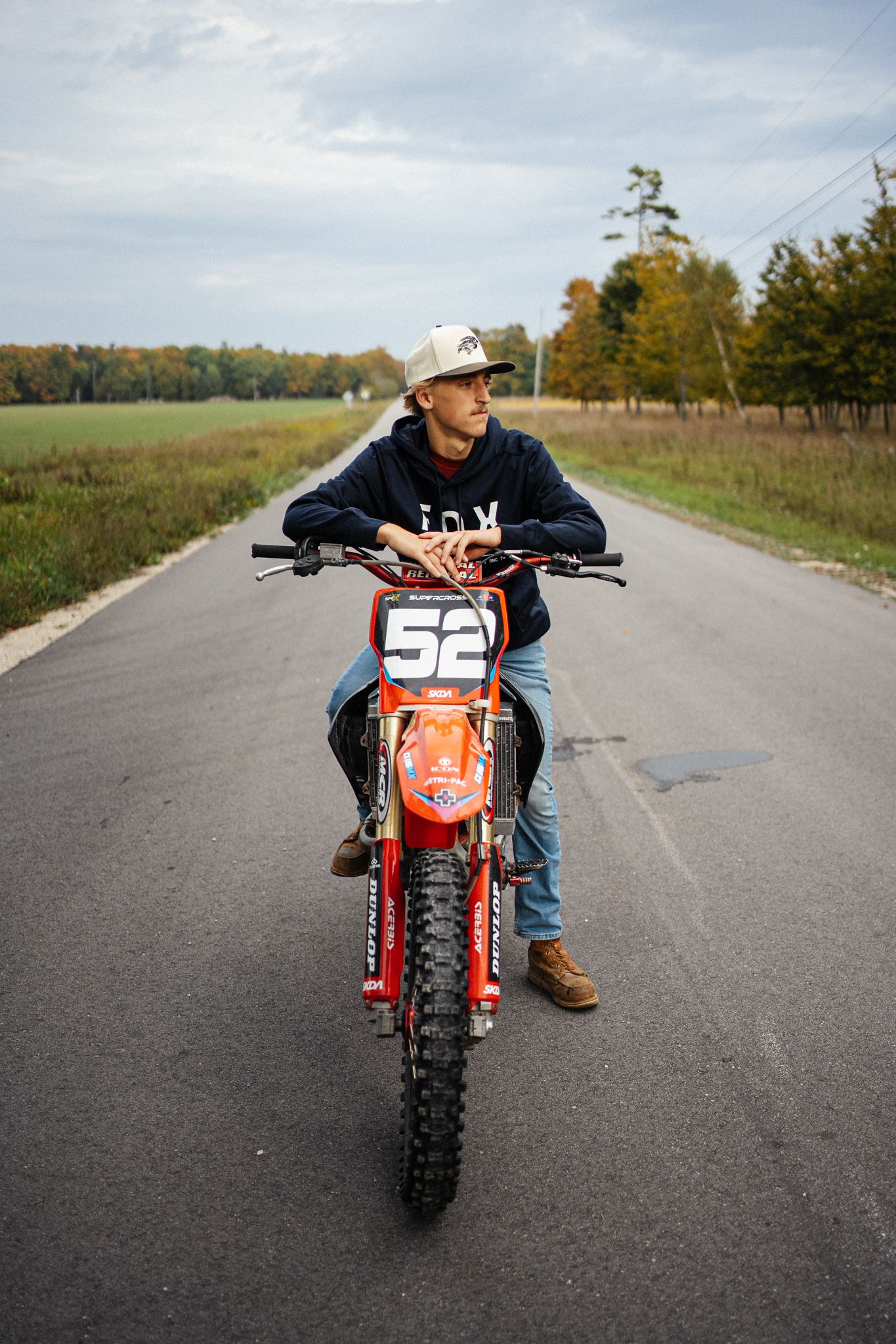 Young man sitting on a red dirt bike numbered 52 on a rural road in autumn.