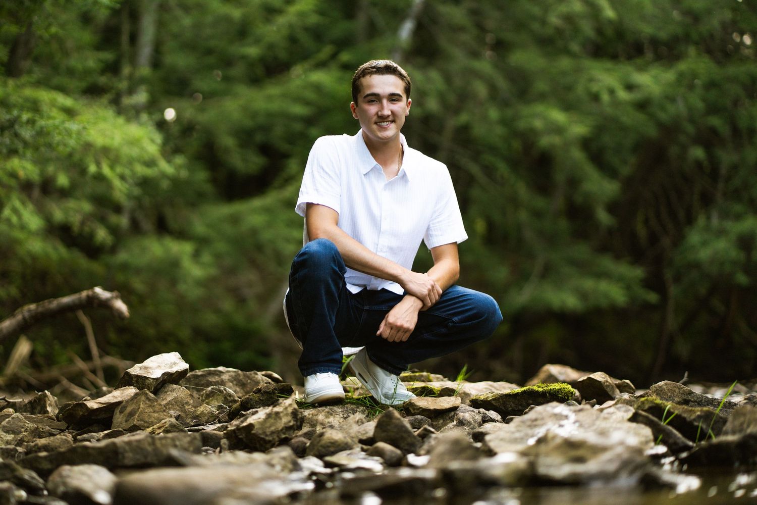 Young man in white polo crouching on rocky creek bed surrounded by lush green forest.