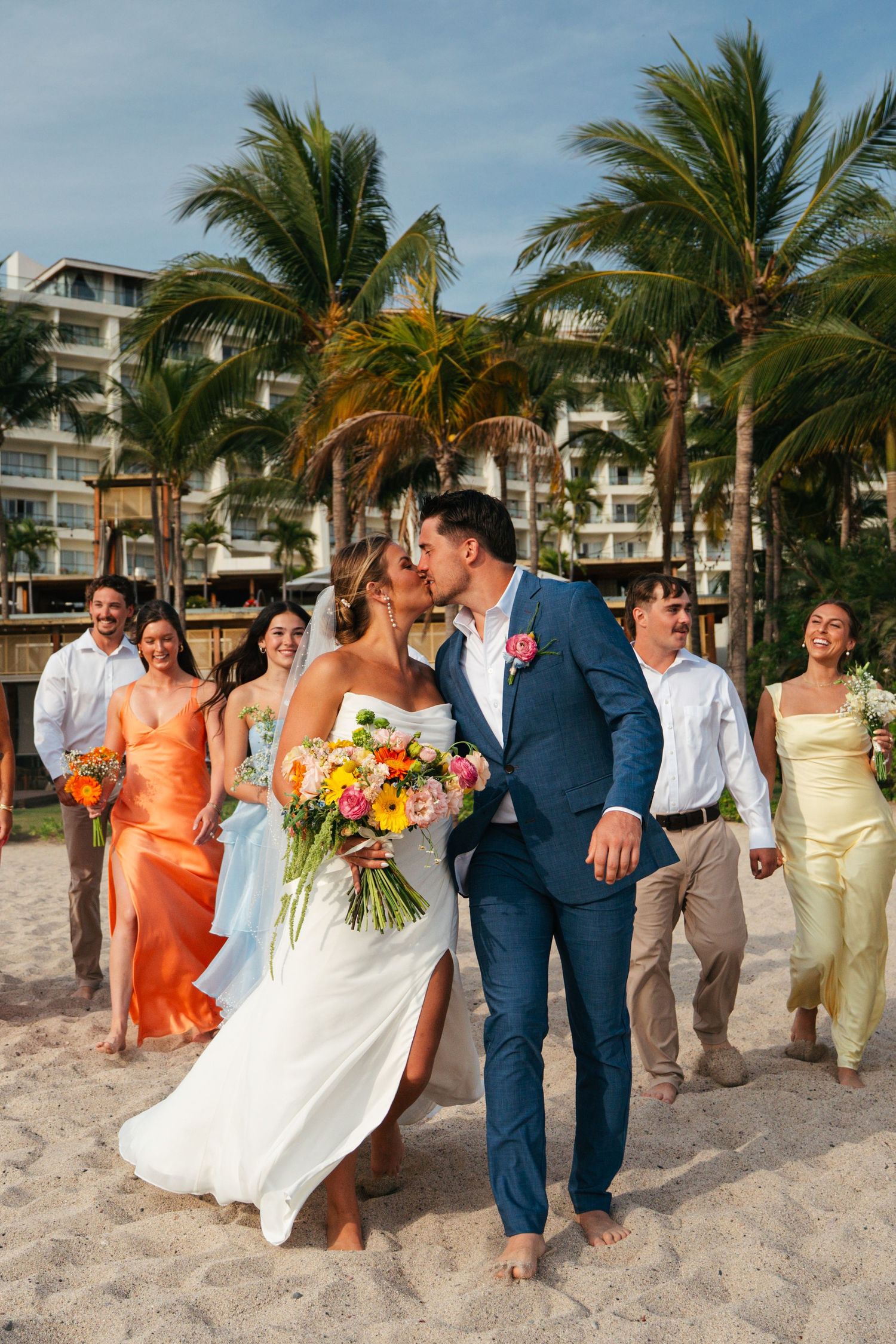 Bride and groom kiss on sandy beach with wedding party and palm trees in background.