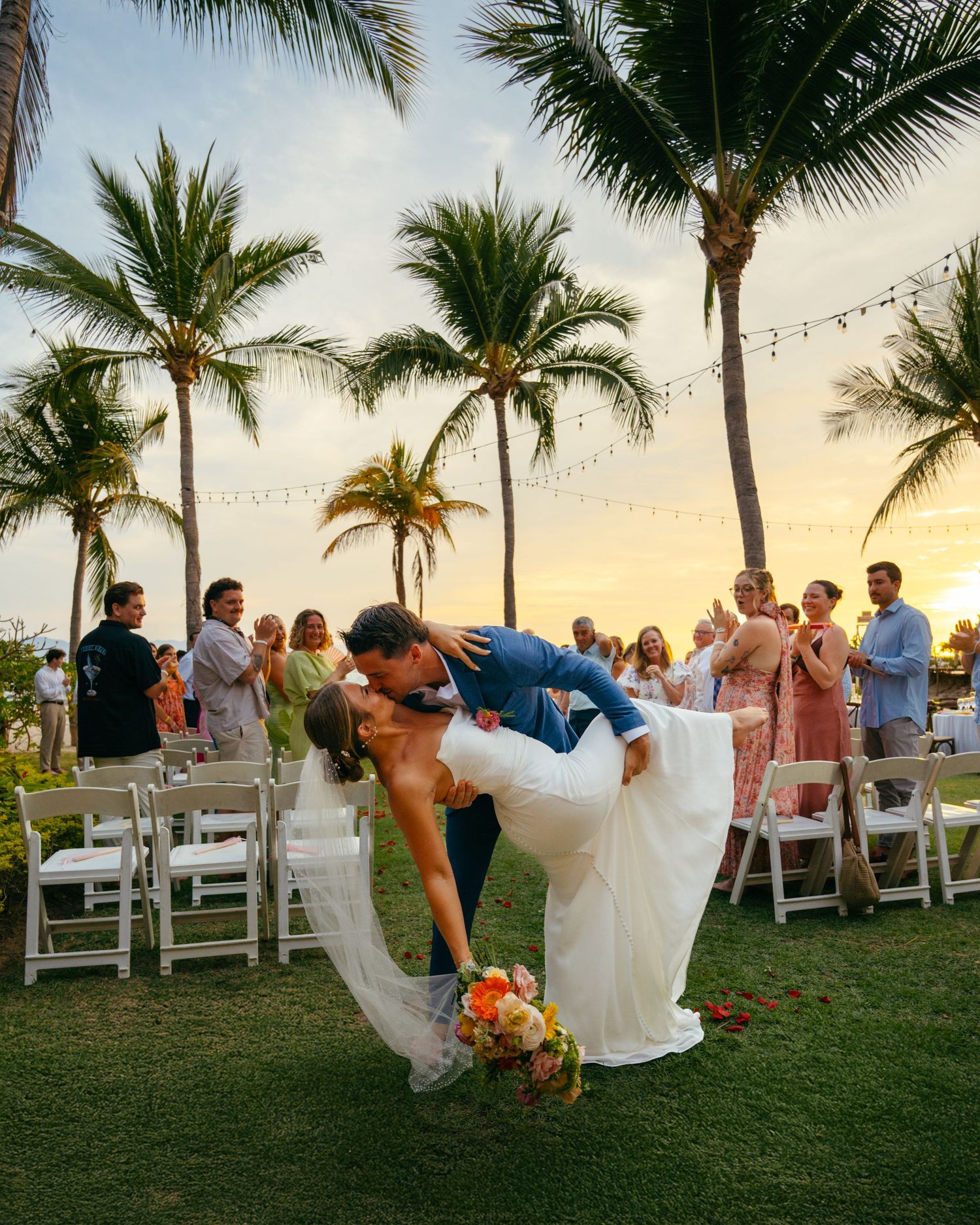 Groom dips bride in a romantic kiss at a tropical beach wedding with a stunning sunset backdrop.