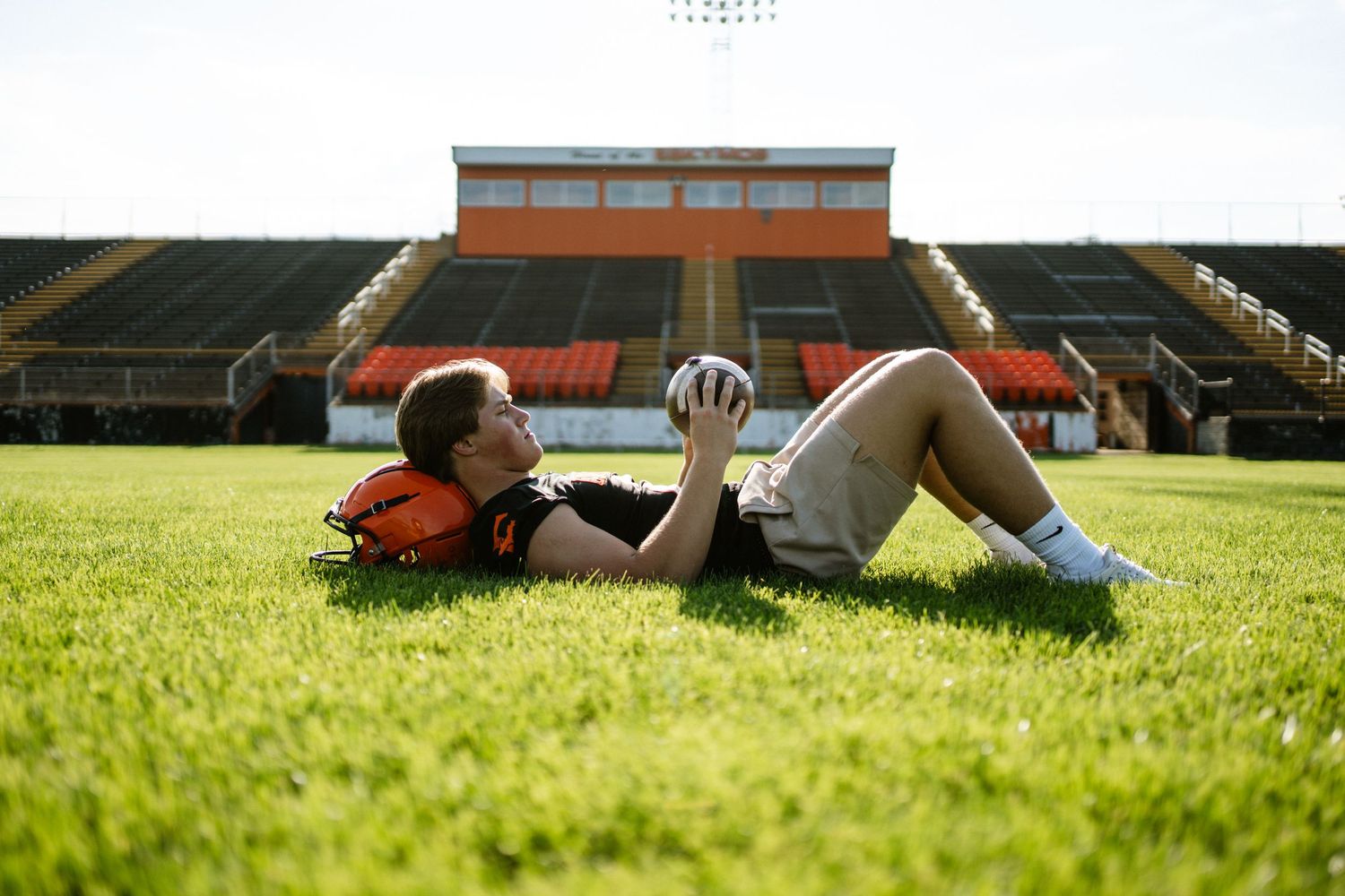 Football player lying on stadium grass holding helmet while gazing up at the sky.