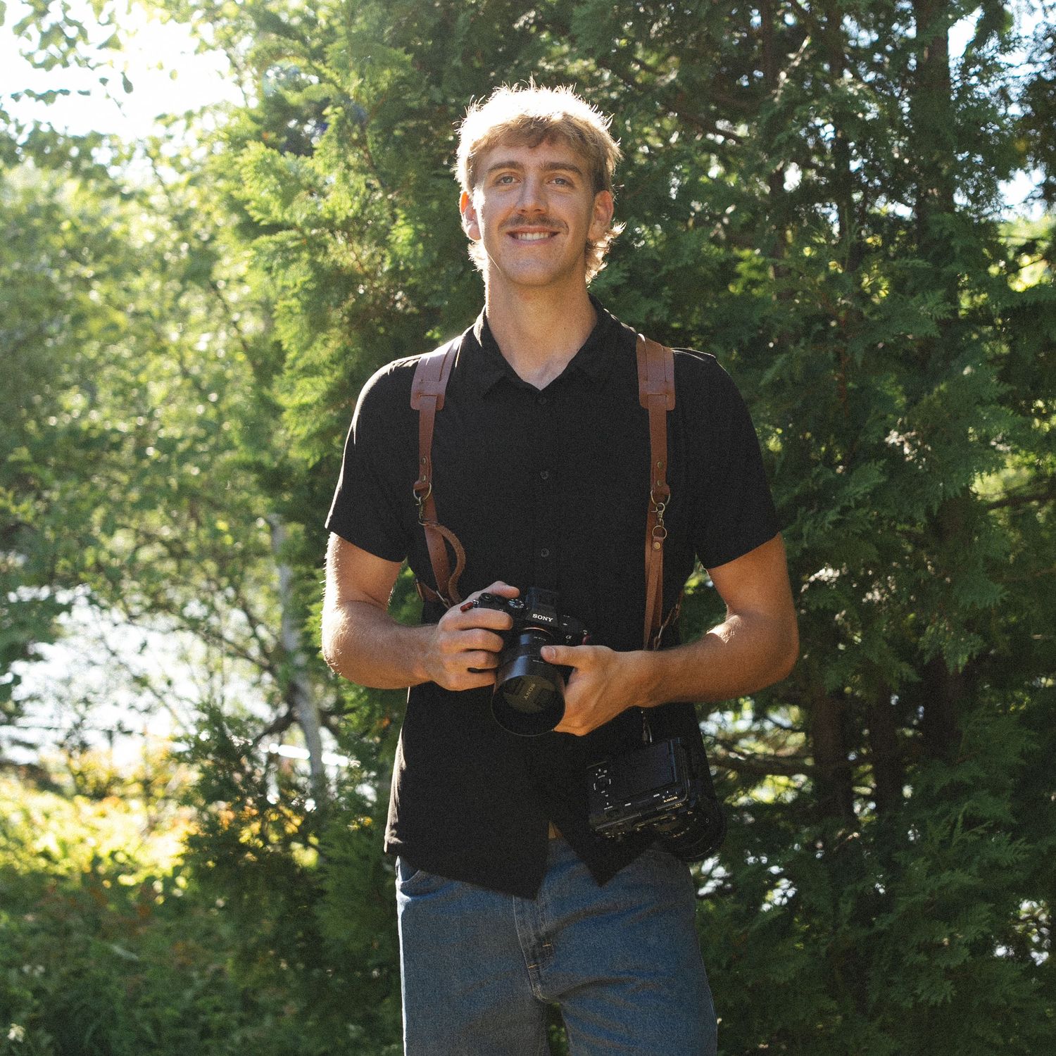 Young man with mustache smiling outdoors, wearing black shirt, holding camera with leather strap, green trees background.