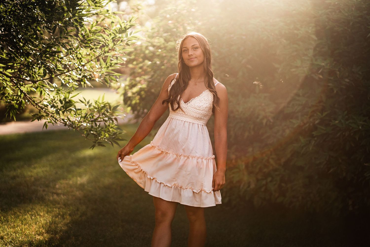 Young woman in white ruffled sundress standing in golden sunlight among green trees.