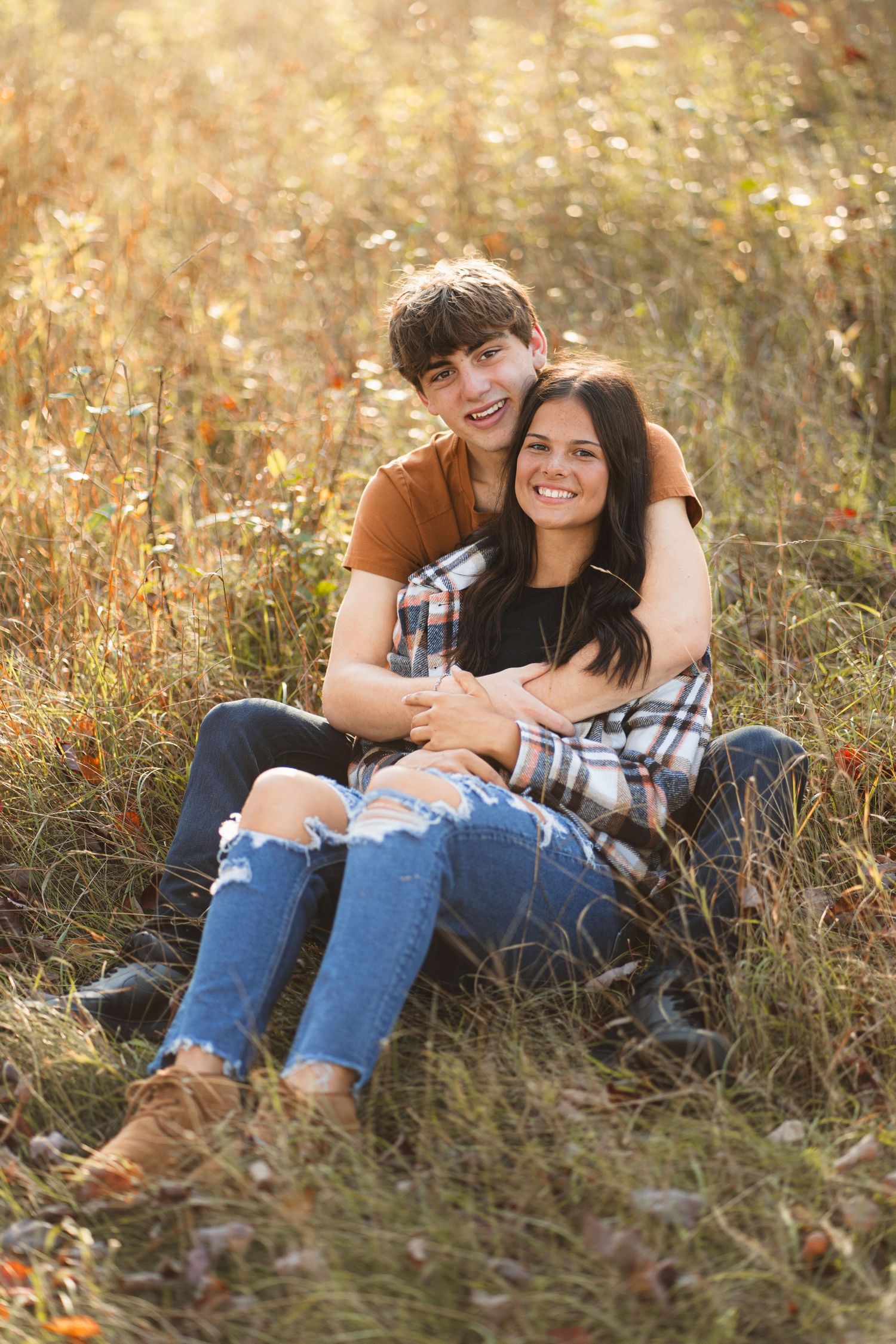 Happy teenage couple sitting together in sunlit golden meadow grass, embracing warmly.