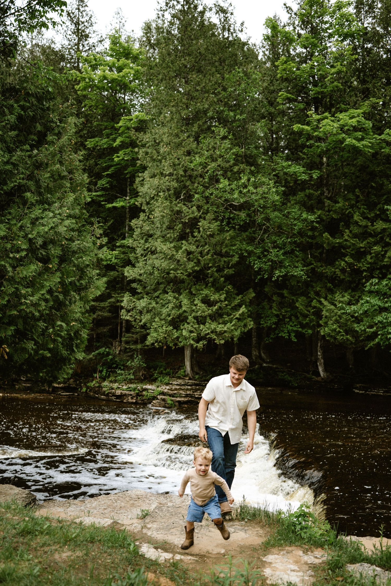 A father and toddler run along a rocky riverbank with a waterfall and lush green forest in the background.