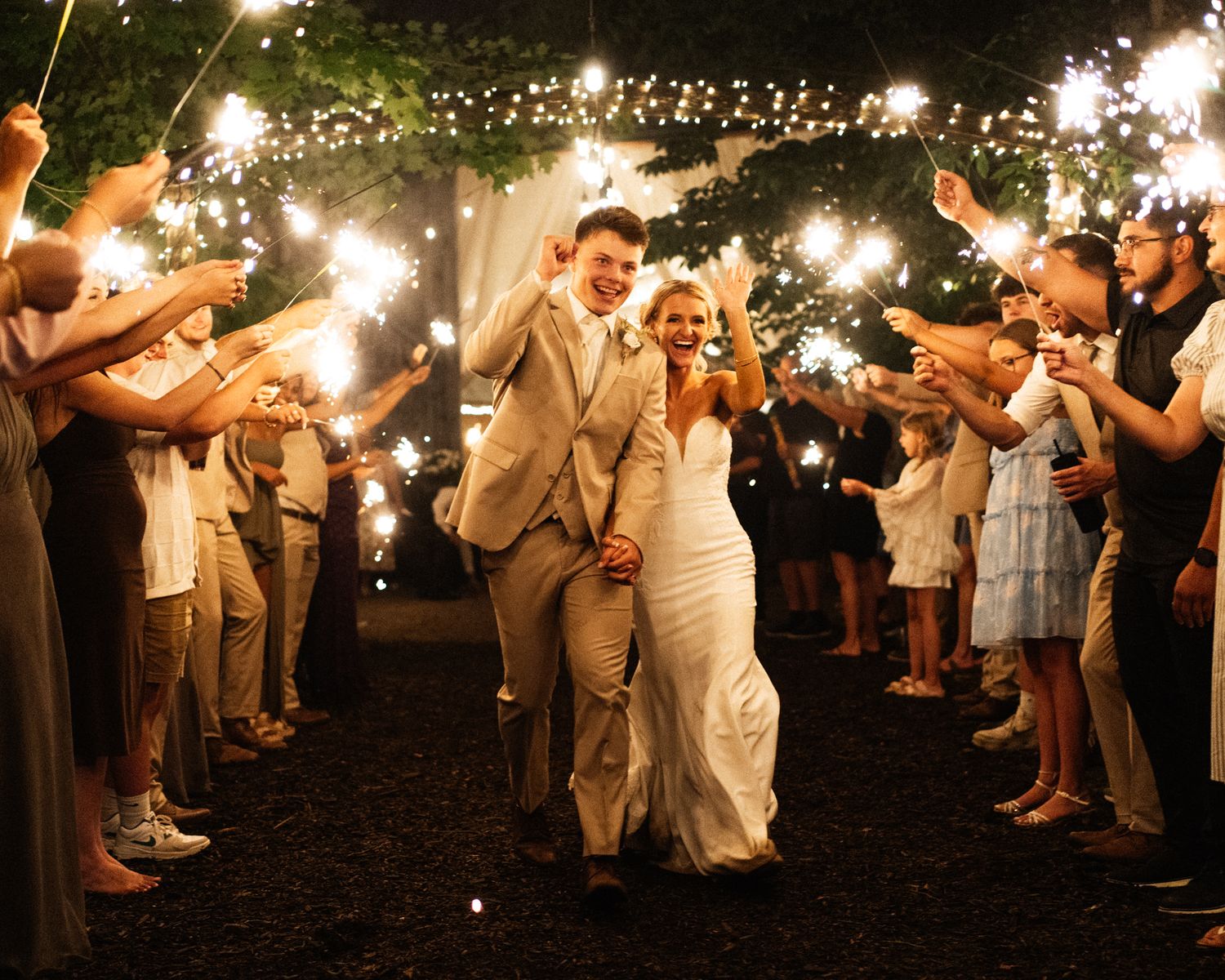 Bride and groom walking through a sparkler tunnel surrounded by cheering wedding guests at night.