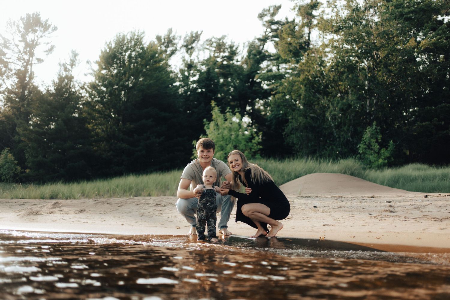 Family of three smiling together on a sandy beach near water, surrounded by lush green trees.