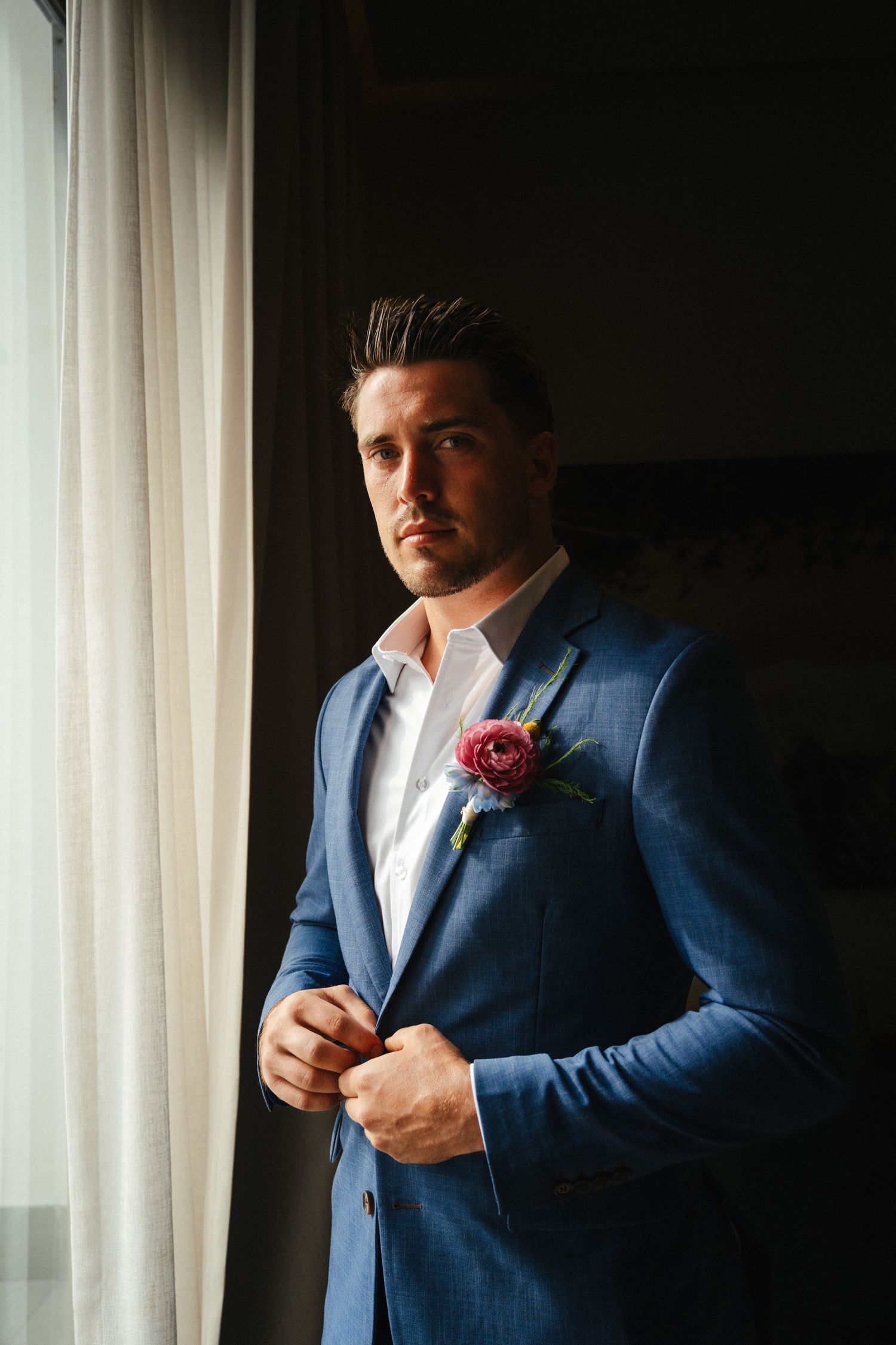 Handsome groom in blue suit with pink boutonniere stands by window in dramatic natural light.