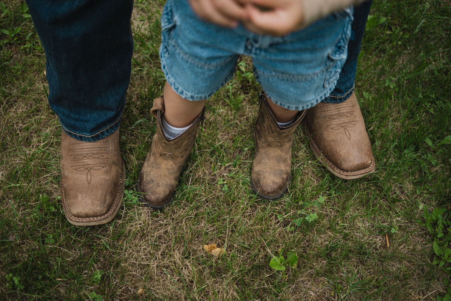 Adult and child wearing matching muddy brown cowboy boots standing on green grass.