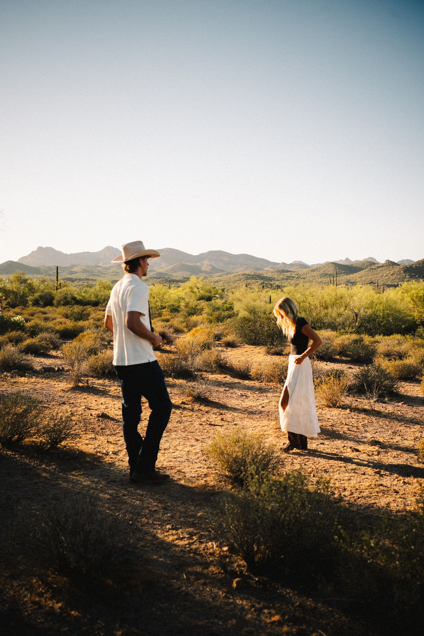 Couple standing in a sunlit desert landscape with mountains in the background during golden hour.