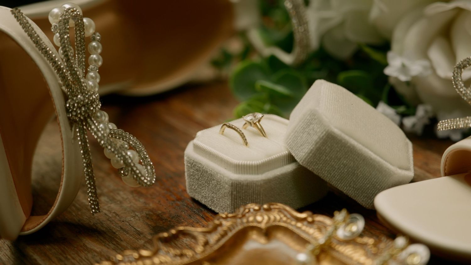 Wedding rings displayed on a velvet box beside pearl-adorned bridal shoes and floral decorations.