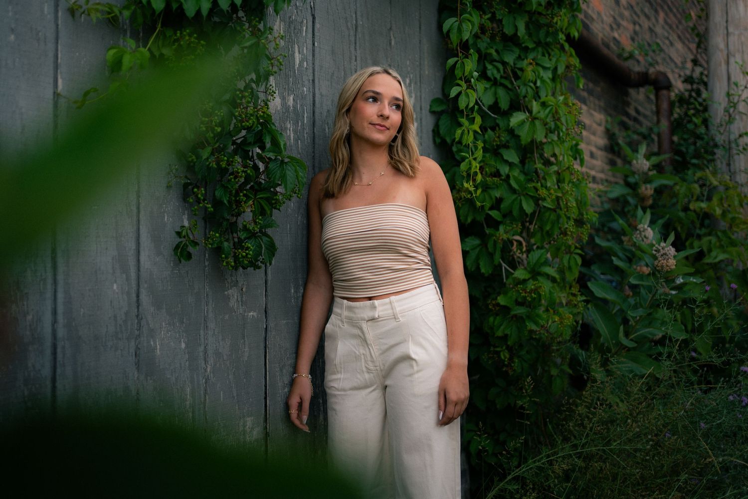 Blonde young woman in beige outfit leaning against a rustic wooden wall with green ivy.