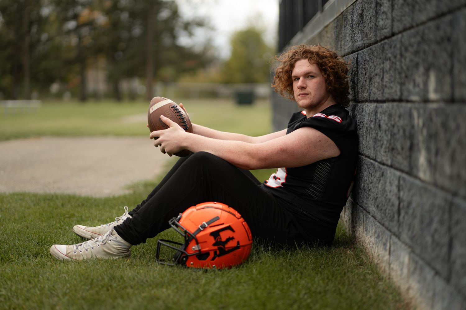 High school football player in black jersey sitting against a wall with helmet and ball.