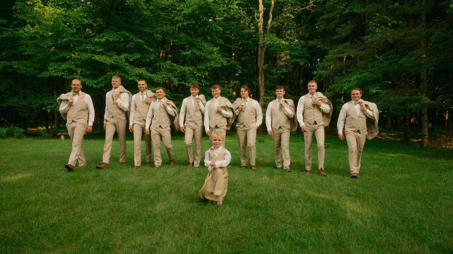 A groom and his groomsmen in tan suits walk playfully across a lush green lawn in a forest setting.