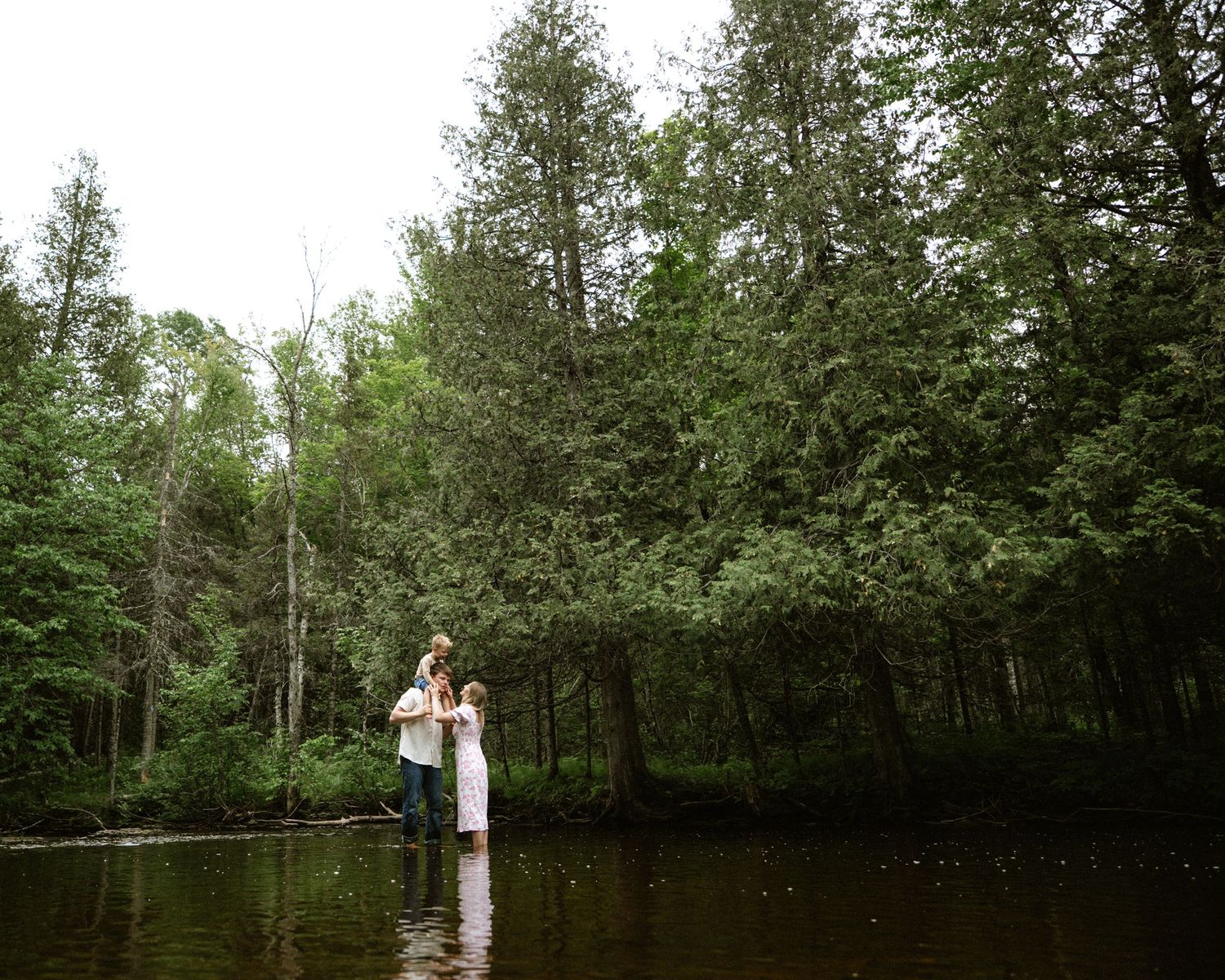 Couple standing in a serene forest river surrounded by tall green trees during an outdoor engagement session.