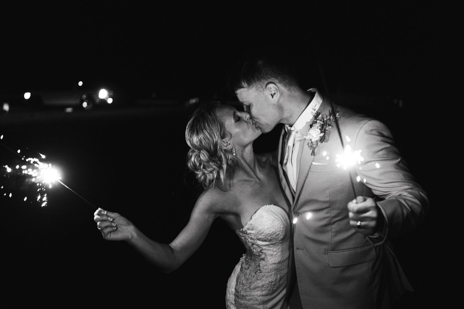 Black and white photo of couple kissing while holding sparklers at night wedding celebration.