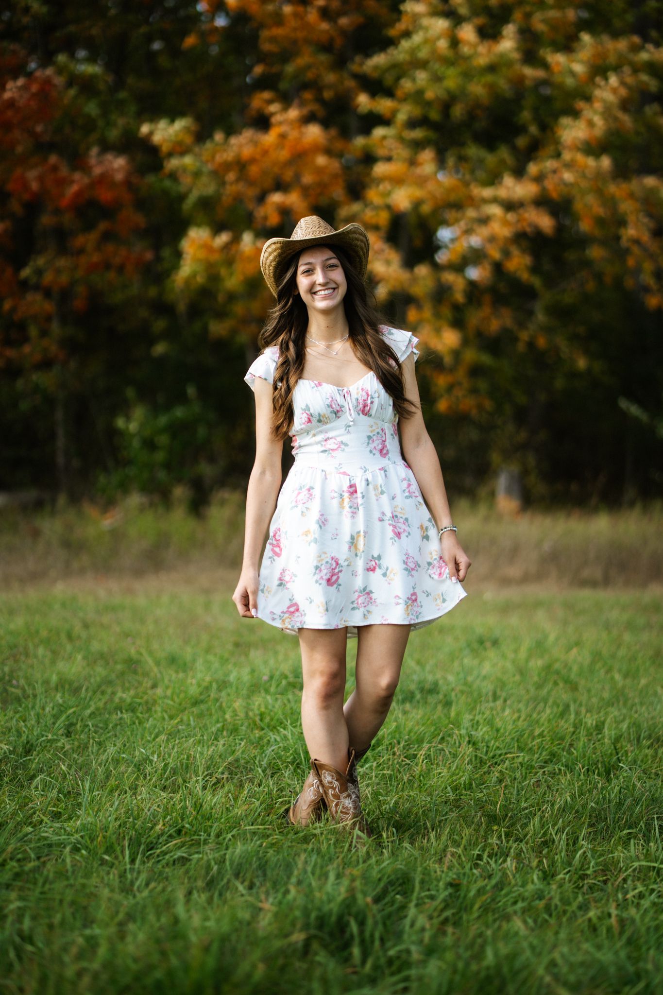 Smiling young woman in floral dress and cowboy hat stands in green field with autumn trees.
