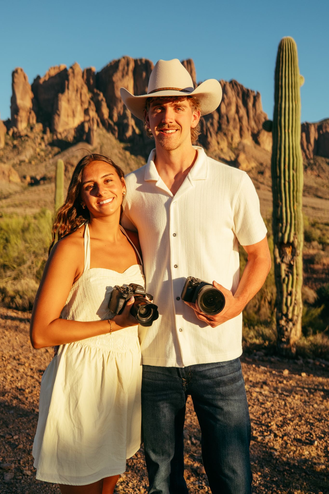 A smiling couple in white outfits holds cameras in an Arizona desert with saguaro cacti and rocky mountains at golden hour.