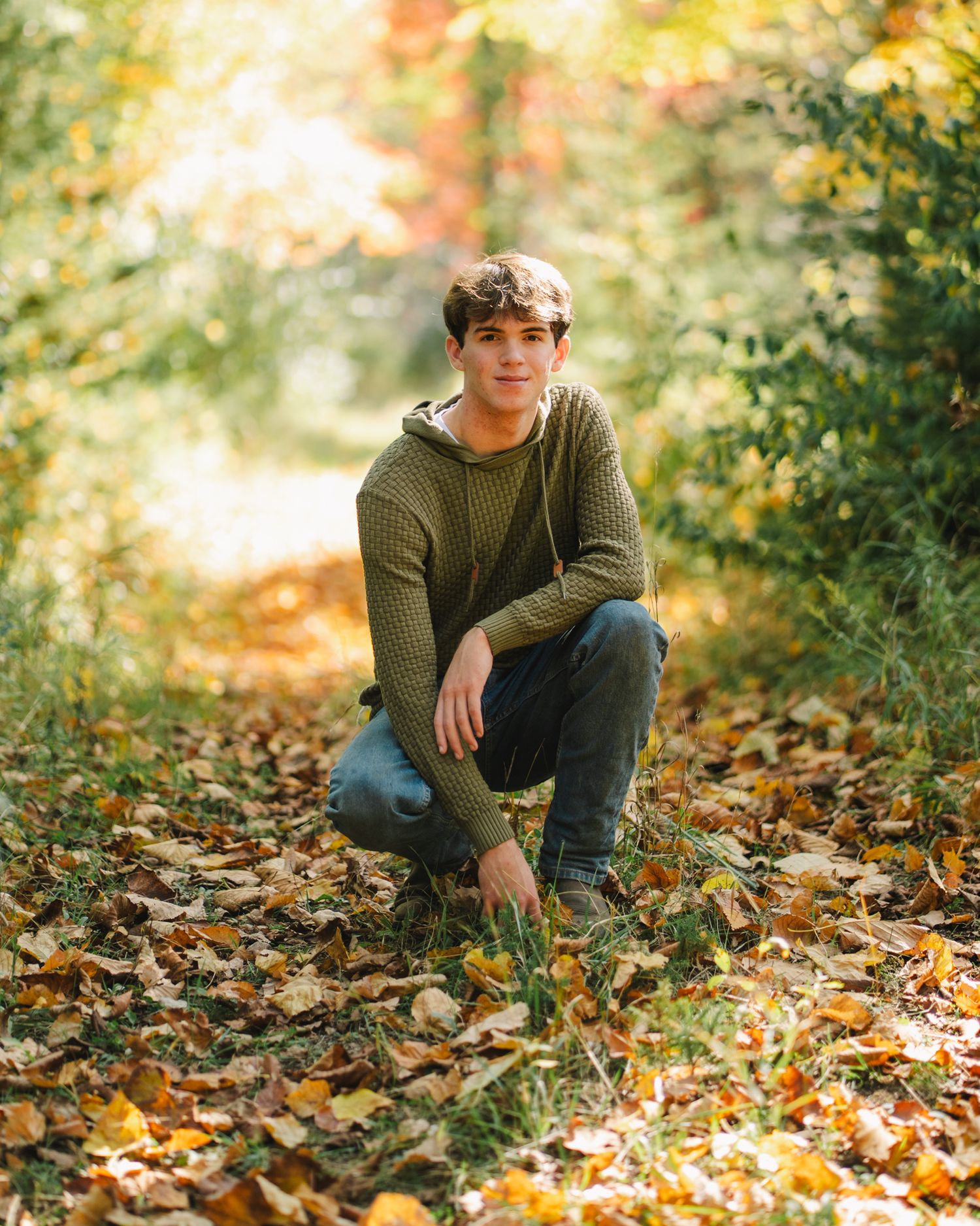 Teen boy in green sweater crouching on fallen autumn leaves in a sunlit forest path.