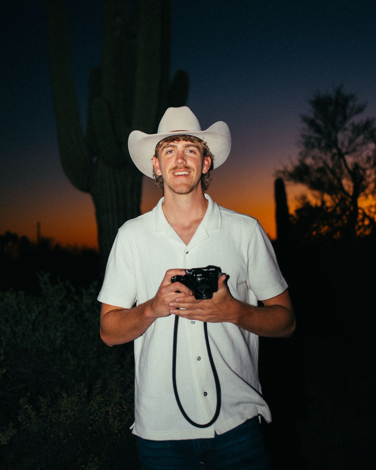Smiling photographer in a white cowboy hat holding a camera at sunset near a saguaro cactus in Arizona.