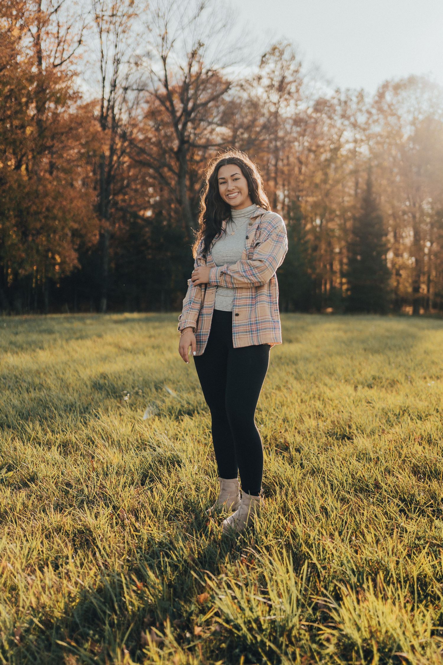 Smiling young woman in plaid jacket and black leggings standing in a sunny autumn field.