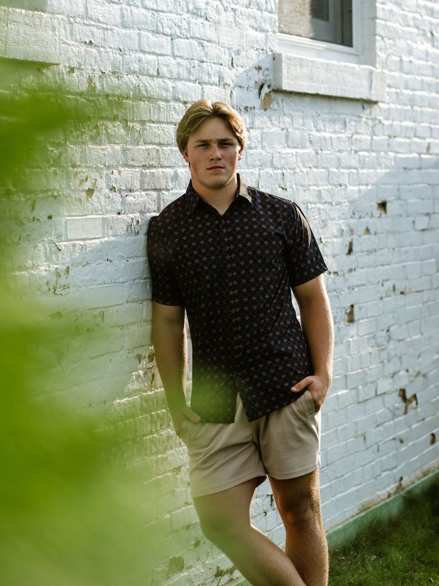 Young blond man in black patterned shirt and khaki shorts leaning against white brick wall outdoors.