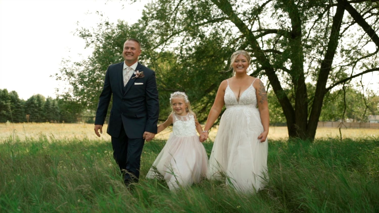Bride and groom walking hand in hand with a young flower girl through a lush green meadow.