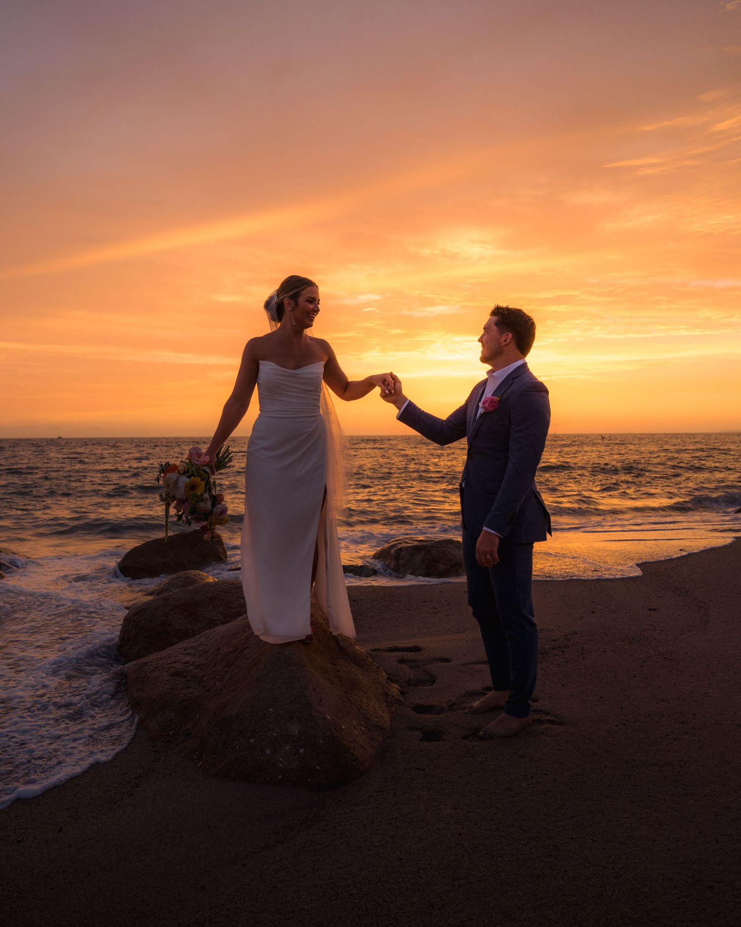 Bride stands on a rock holding groom's hand at a breathtaking ocean sunset wedding photo.